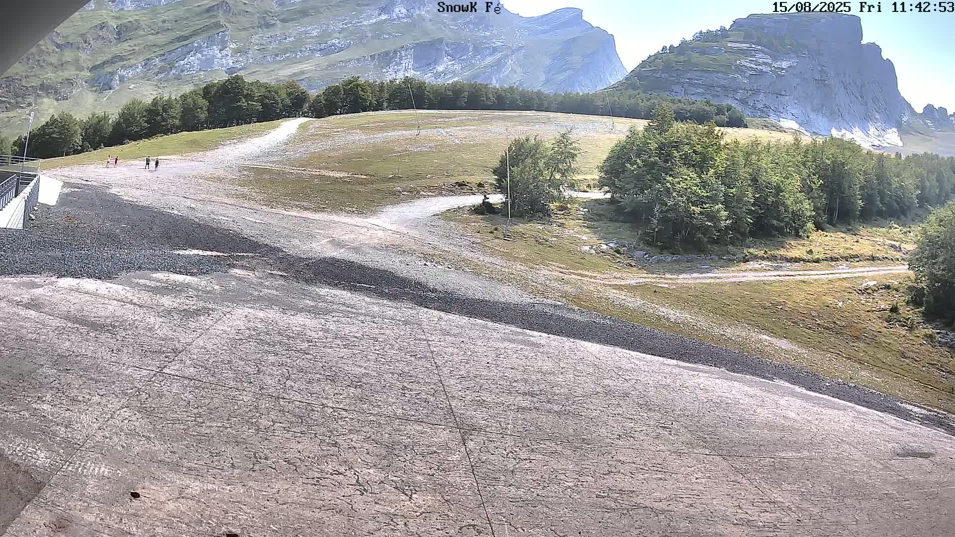 A dry, gravelly slope descends toward a viewer, leading to a grassy area with trees and rocky mountains in the background under a sunny sky.