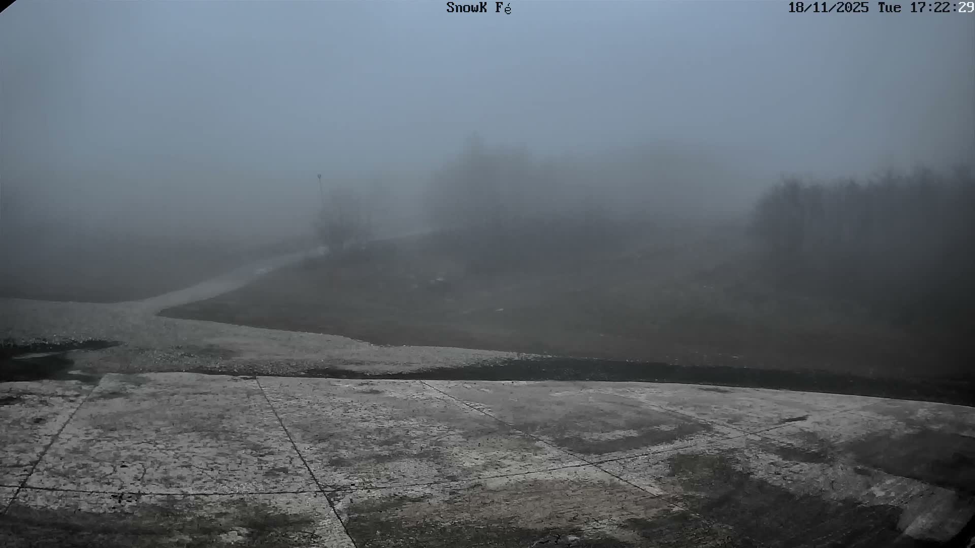 A dry, gravelly slope descends toward a viewer, leading to a grassy area with trees and rocky mountains in the background under a sunny sky.