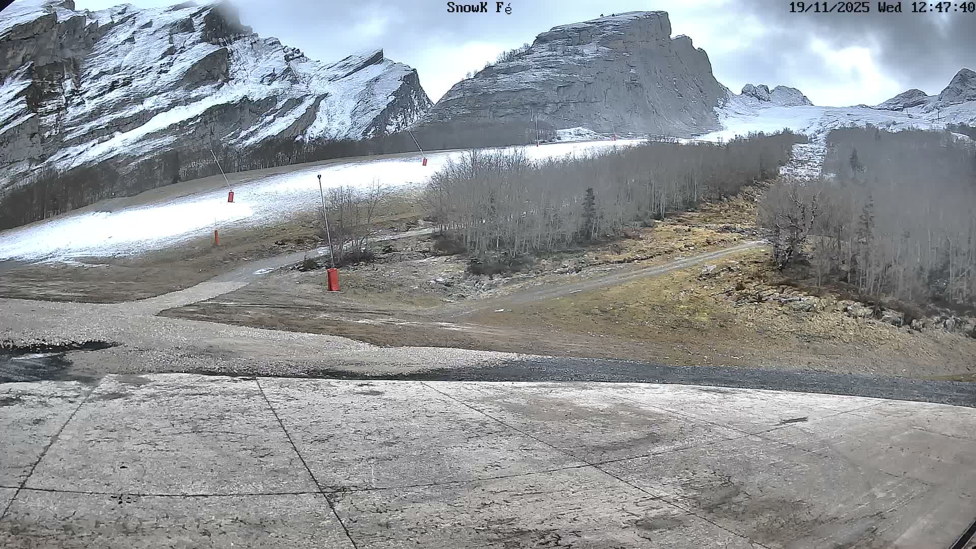 A dry, gravelly slope descends toward a viewer, leading to a grassy area with trees and rocky mountains in the background under a sunny sky.