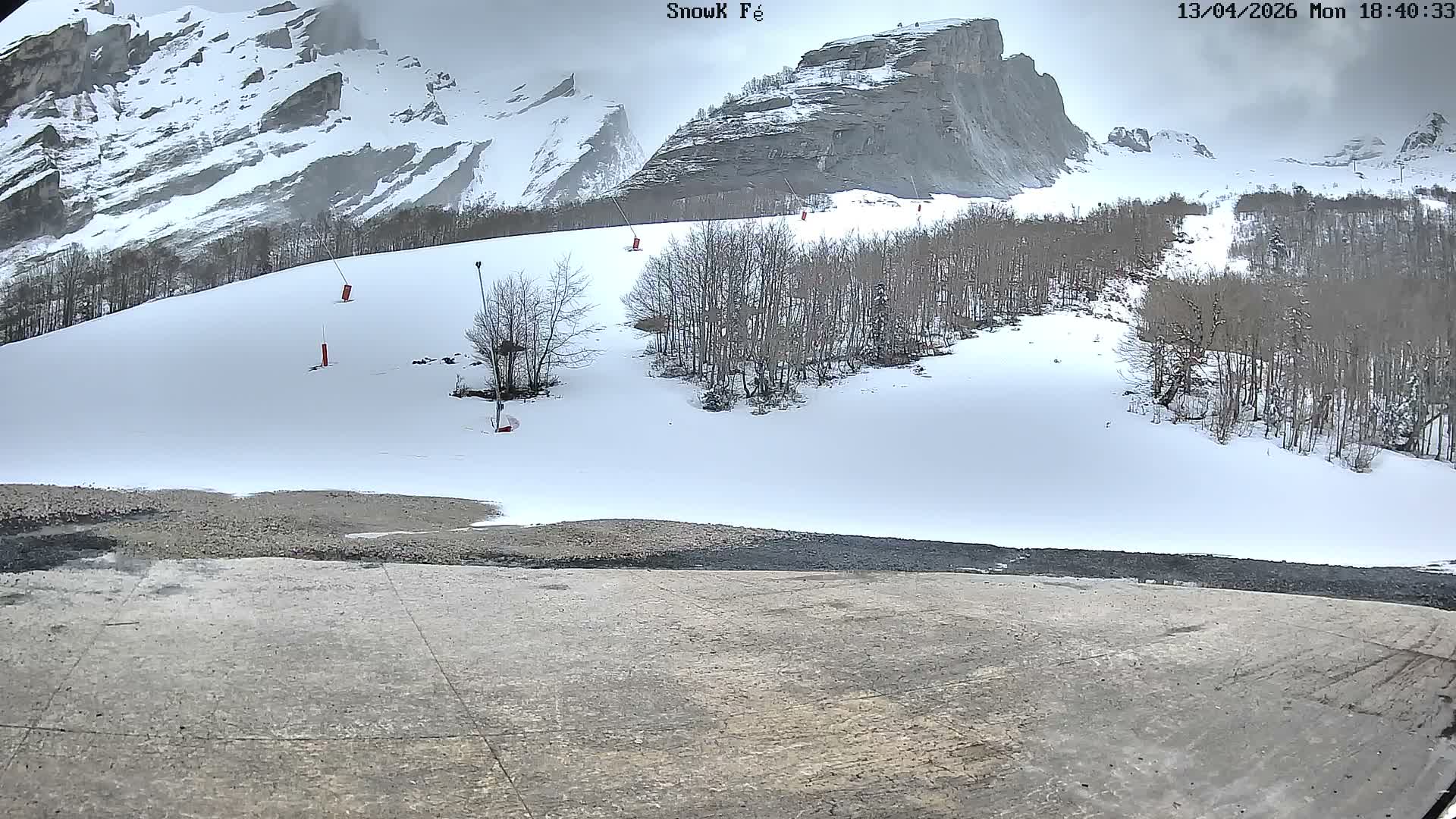 A snow-covered mountain landscape with bare trees, visible ski lift pylons, and a slushy foreground path is presented under an overcast sky.