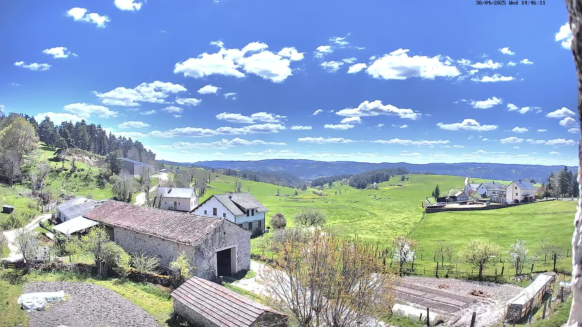 A sunny day with scattered clouds shows a rural landscape with several stone buildings, green fields, and a forest in the background.