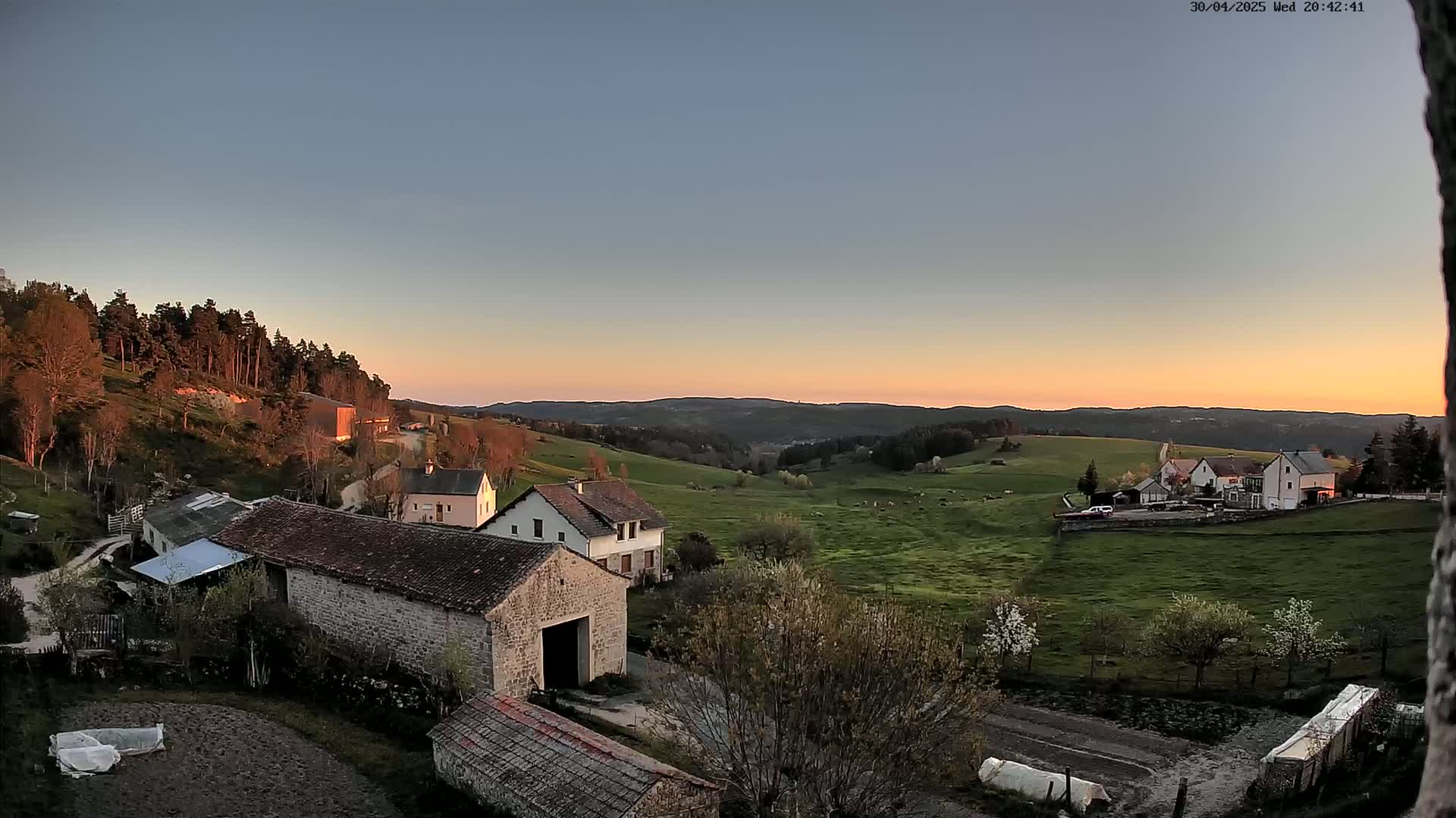 A sunset illuminates a pastoral valley with several farmhouses and barns, surrounded by green fields and a forest in the background.