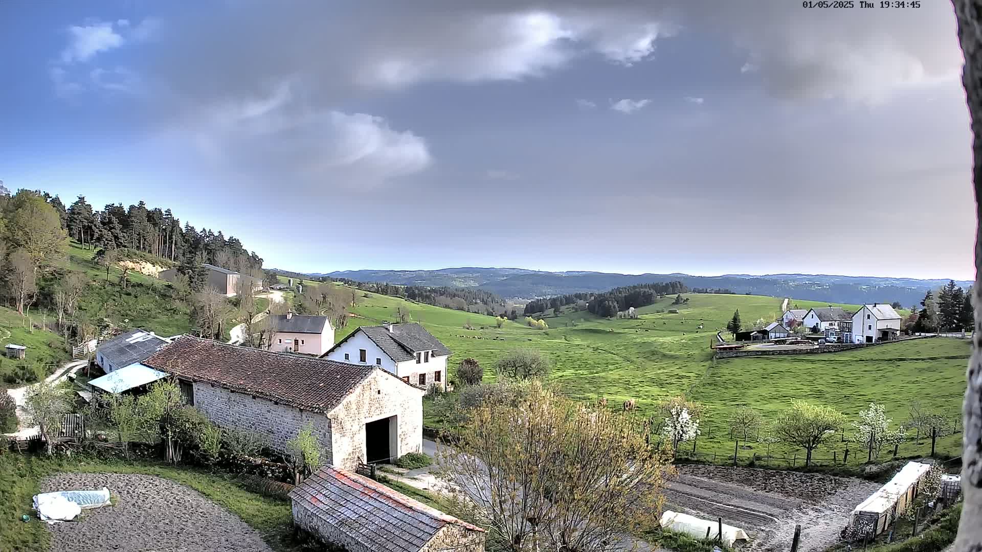 Stone farm buildings sit on a hillside overlooking a valley of green pastureland under a partly cloudy sky.