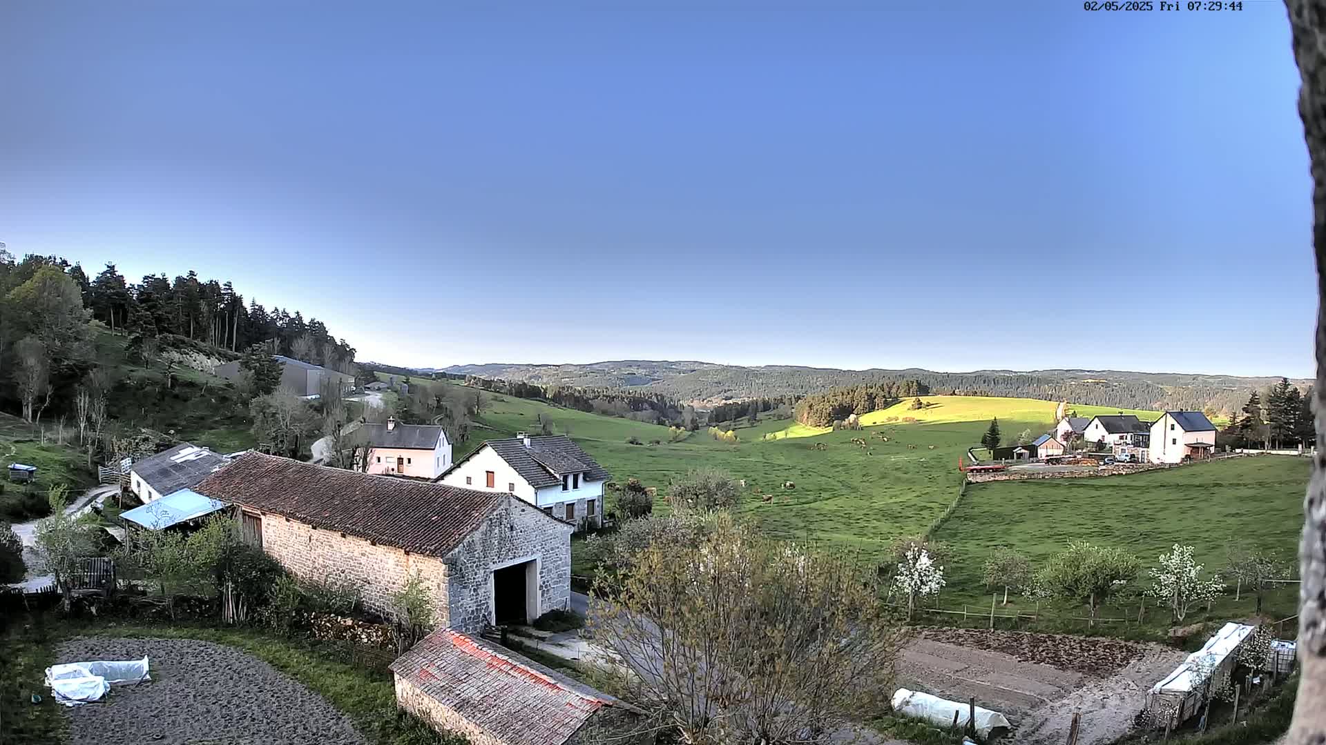A rural scene on a clear, sunny day shows several stone buildings and houses nestled in a green valley, surrounded by fields and a distant forest.