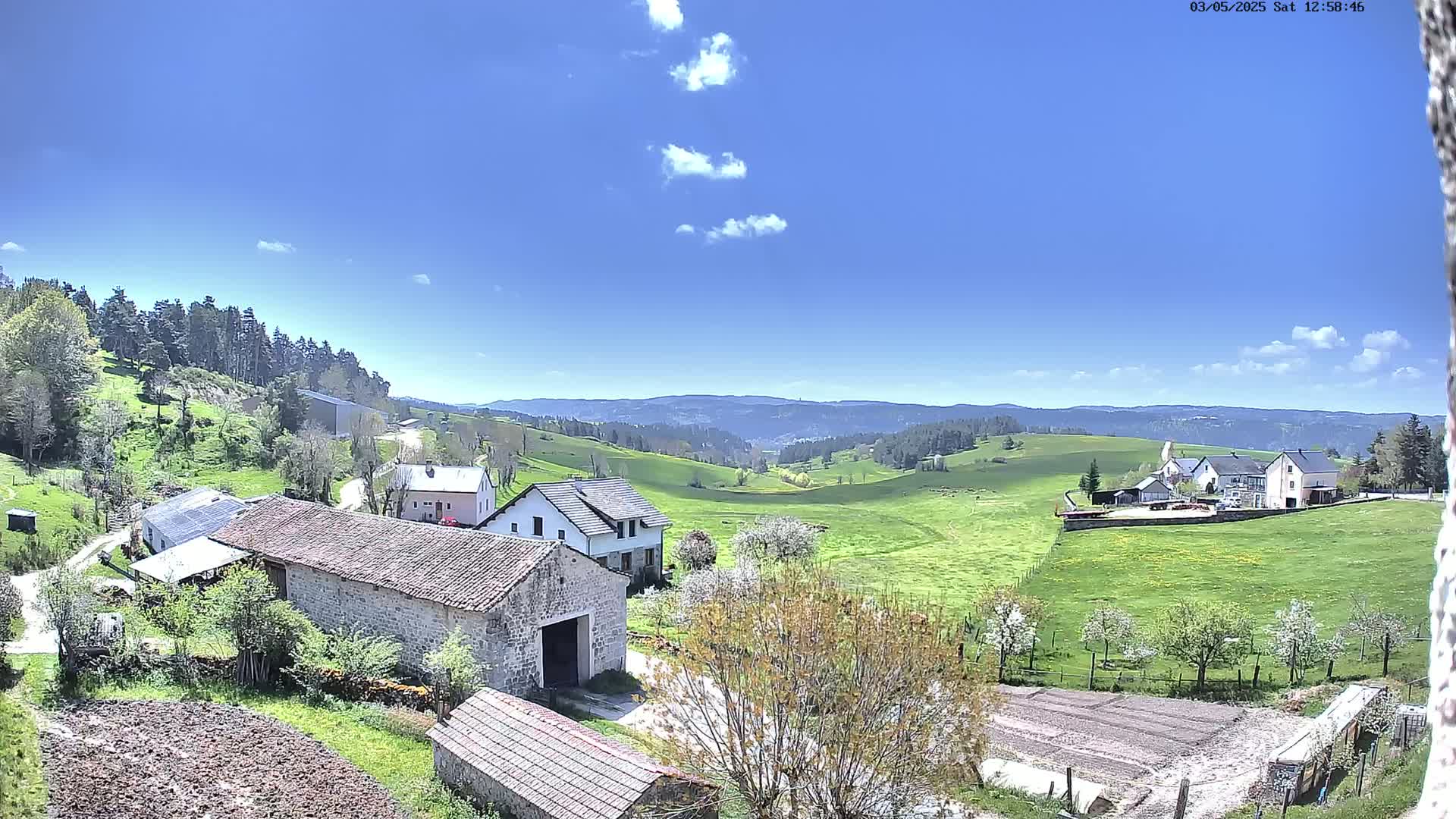 A sunny day reveals a rural landscape with stone buildings, green pastures, and a distant mountain range under a mostly clear blue sky.