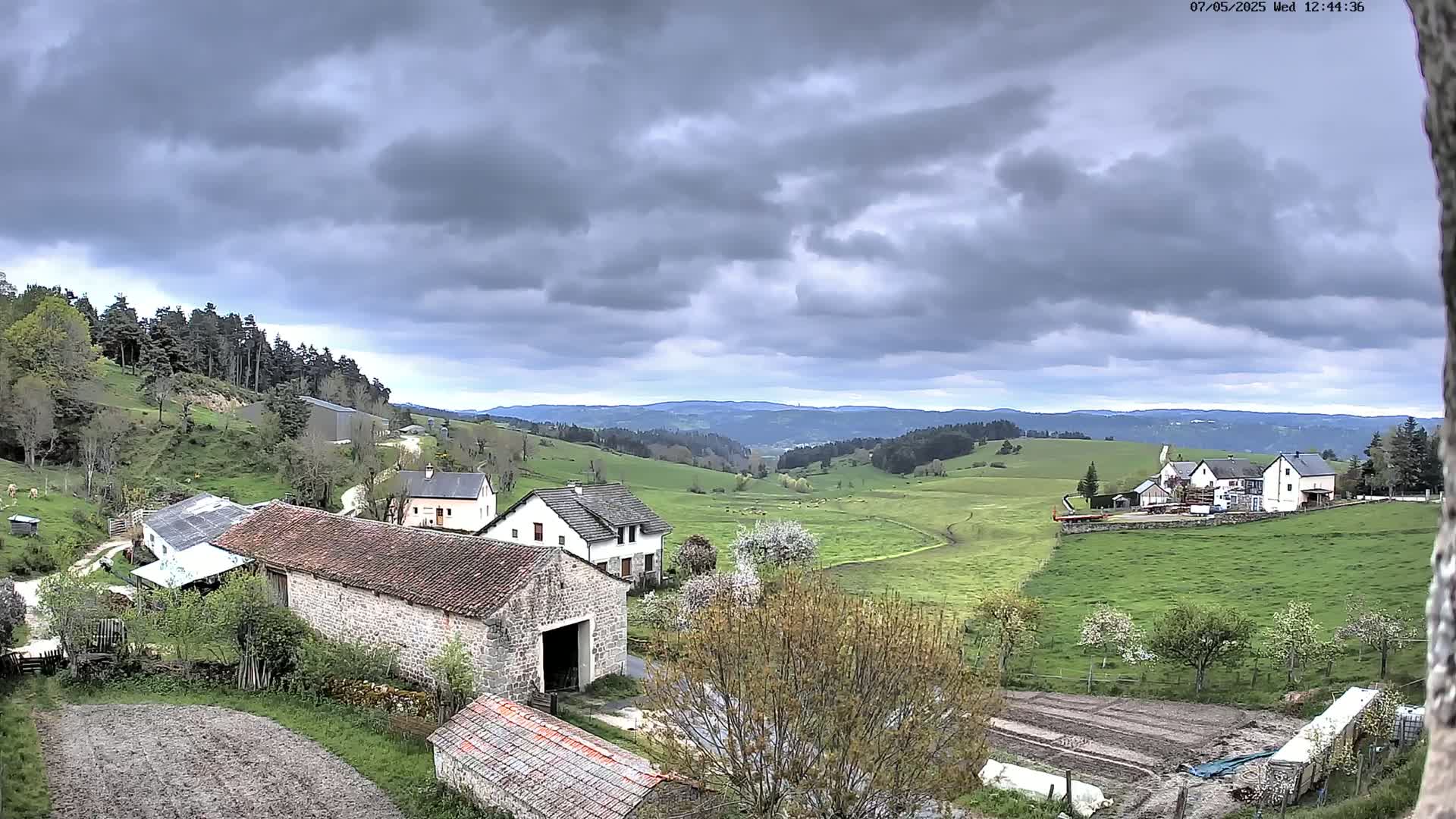 Stone farm buildings sit on a hillside overlooking a sprawling green valley under a cloudy sky.