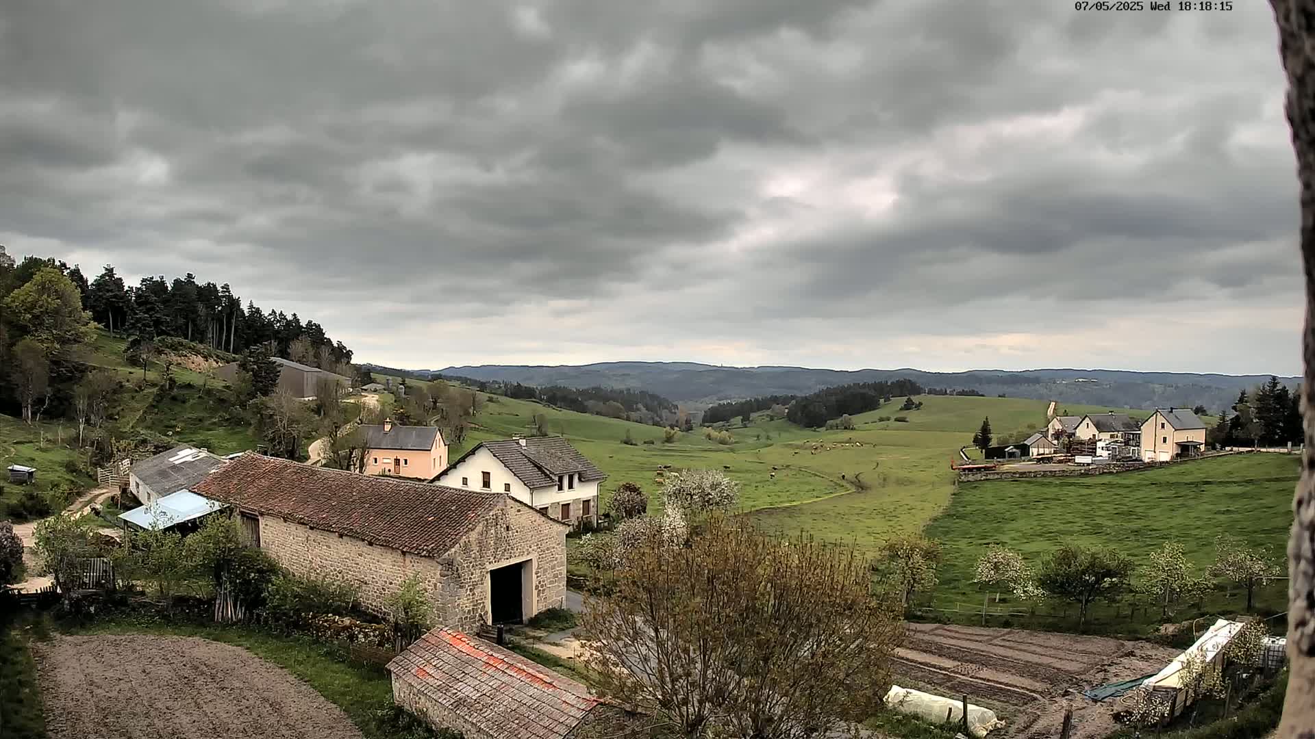 A rural landscape under a cloudy sky shows several stone and stucco farm buildings nestled in a valley surrounded by rolling green hills and dark forest.