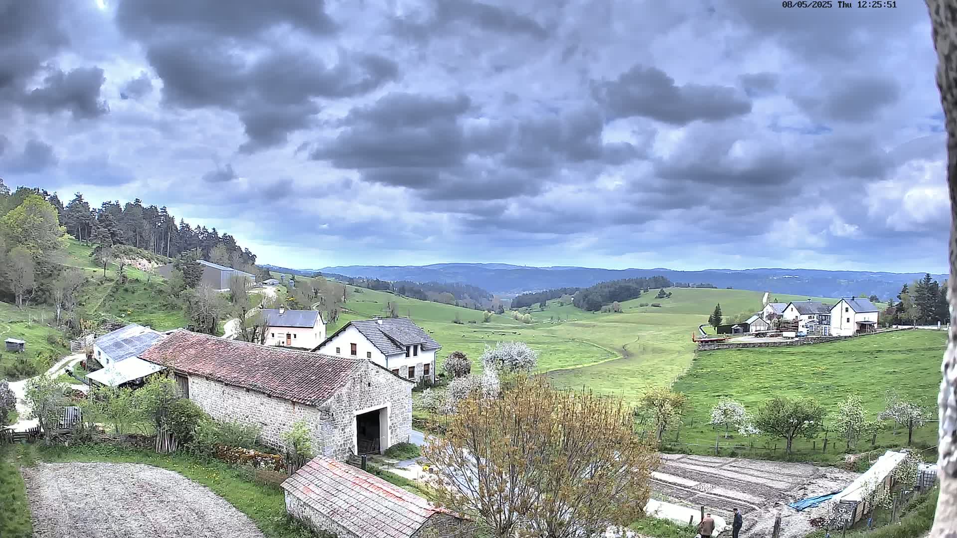 A rural landscape under a cloudy sky shows several stone buildings, green fields, and a distant mountain range.