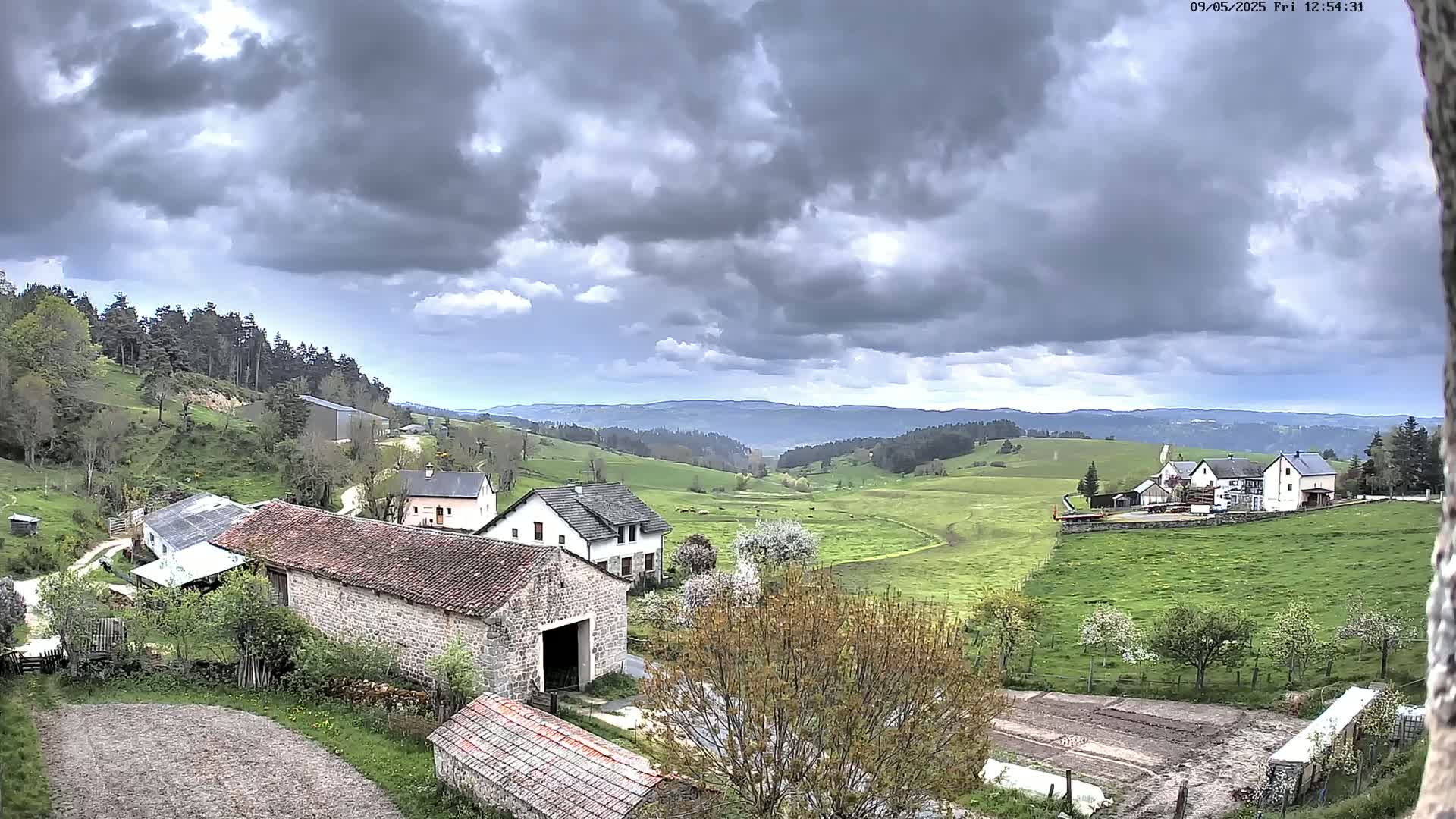 A rural valley with several buildings and farm structures is visible under a cloudy sky.