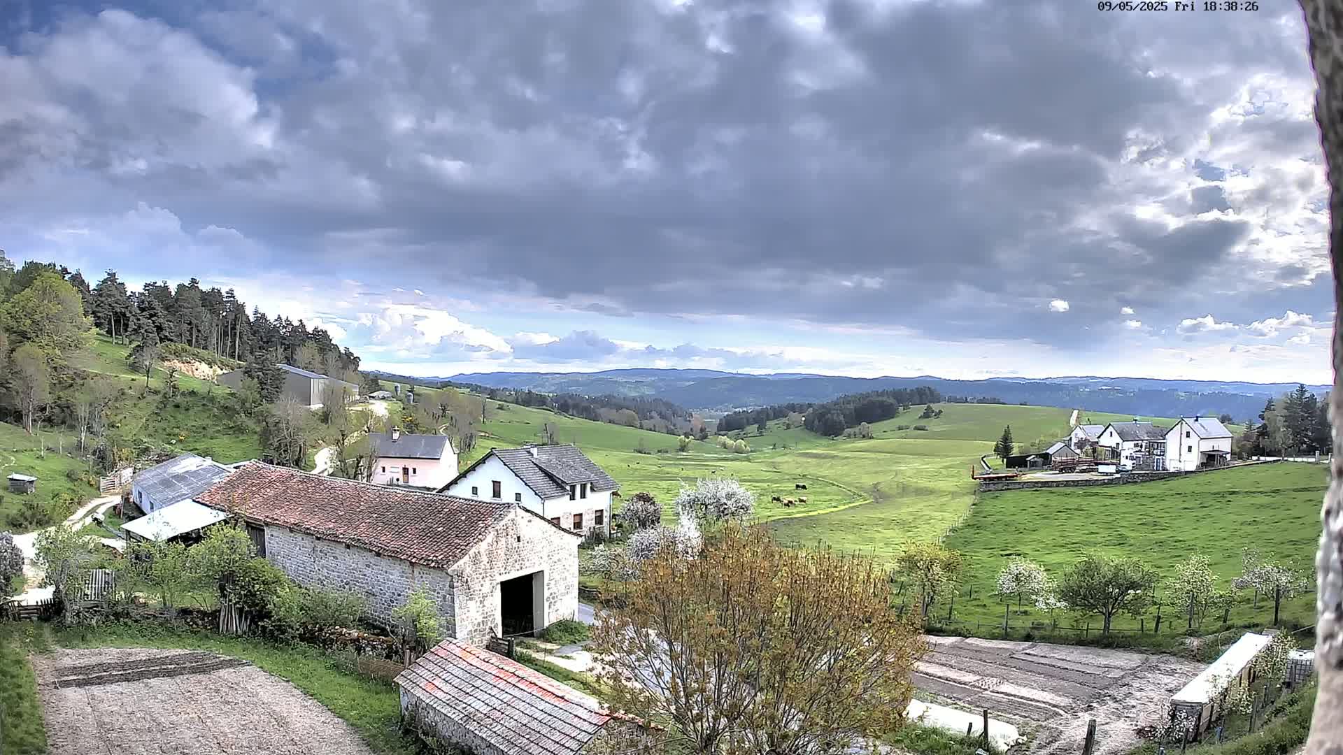 A pastoral valley scene under a partly cloudy sky shows several farm buildings nestled among green hills and fields dotted with trees.