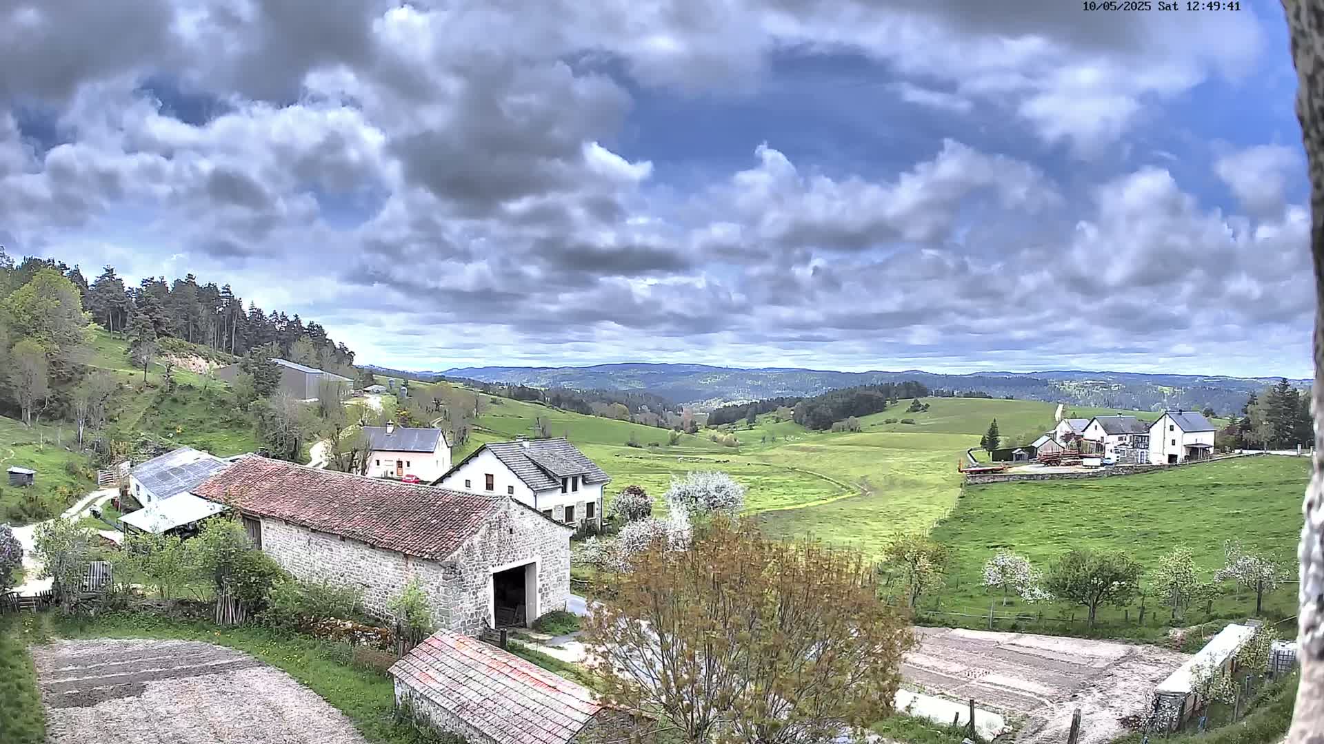 A partly cloudy sky overlooks a rural landscape of stone farm buildings, green pastures, and a distant line of trees under a partly cloudy sky.