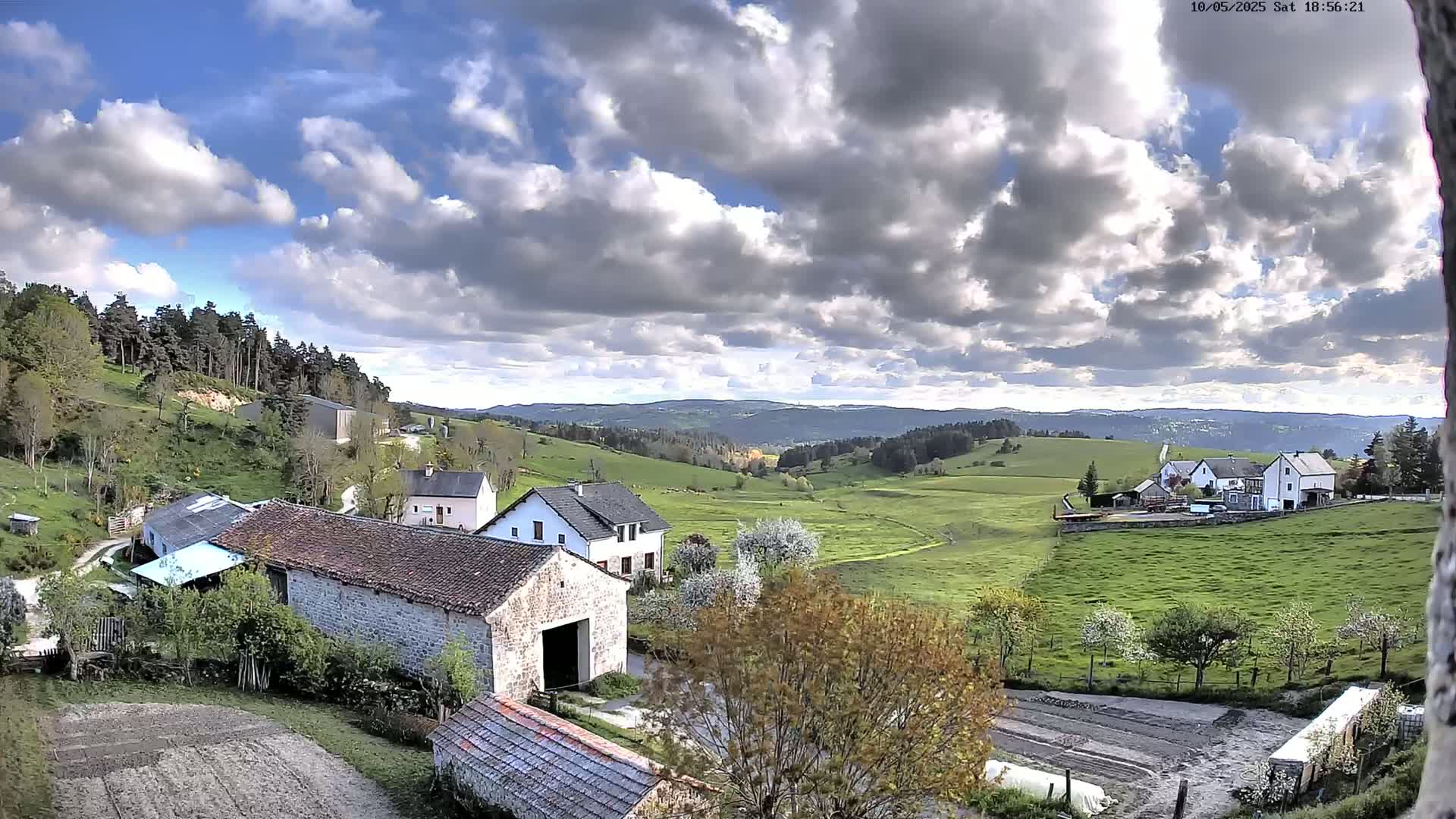 A partly cloudy day reveals a rural landscape with several stone and white buildings nestled among green pastures and a forest-covered hill.