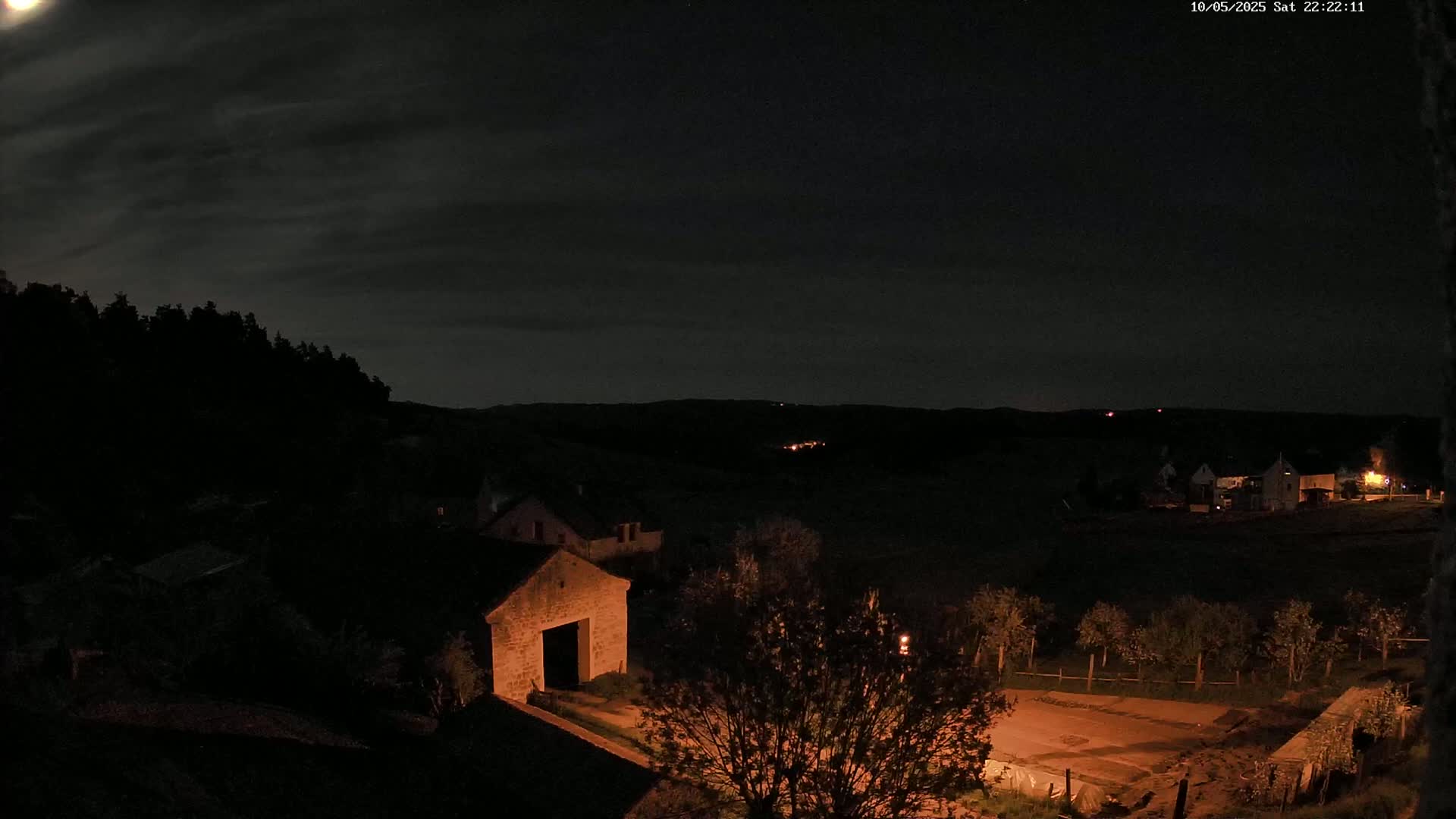 A nighttime view of a rural area shows a stone barn and other buildings under a partly cloudy sky illuminated by a bright moon.