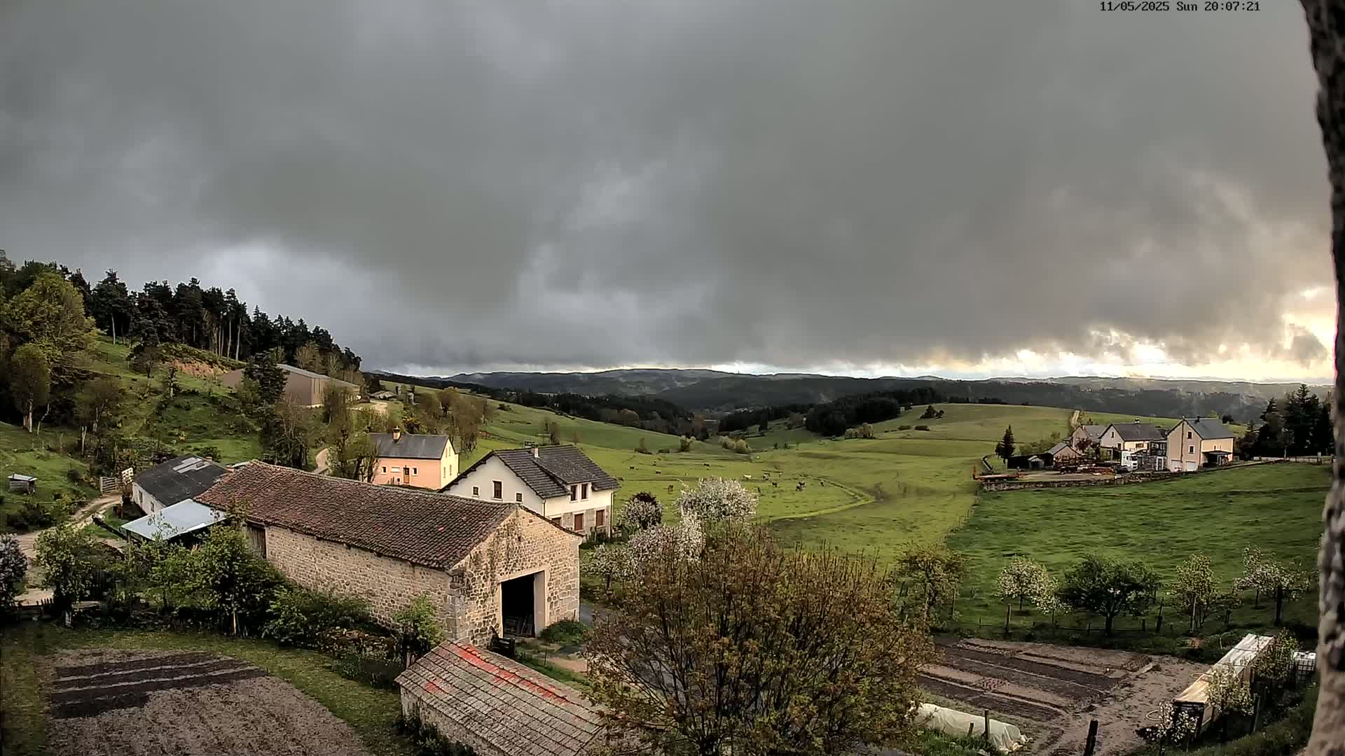 Stone farm buildings and houses sit on a verdant hillside under a heavily clouded sky.