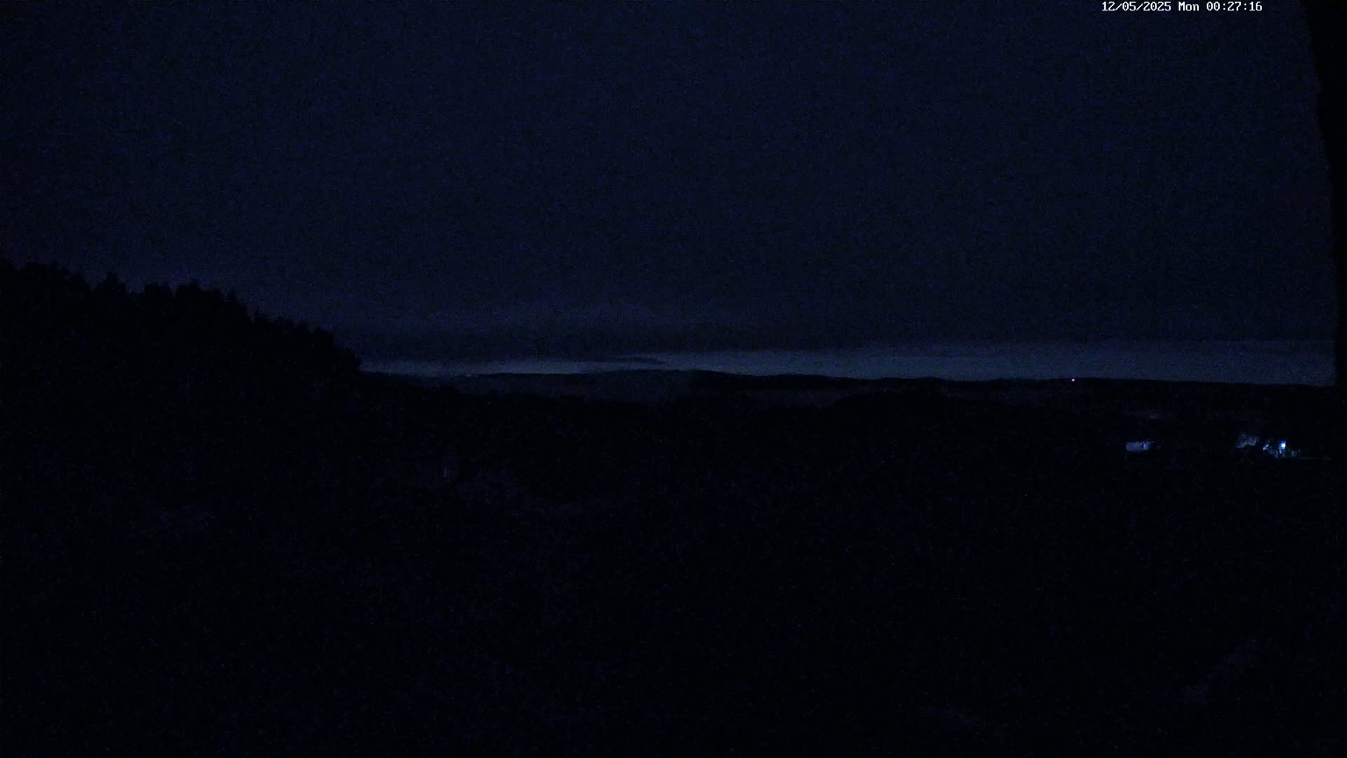 A dark, nighttime landscape shows a silhouetted line of trees and distant low-lying clouds under a mostly clear, dark sky.