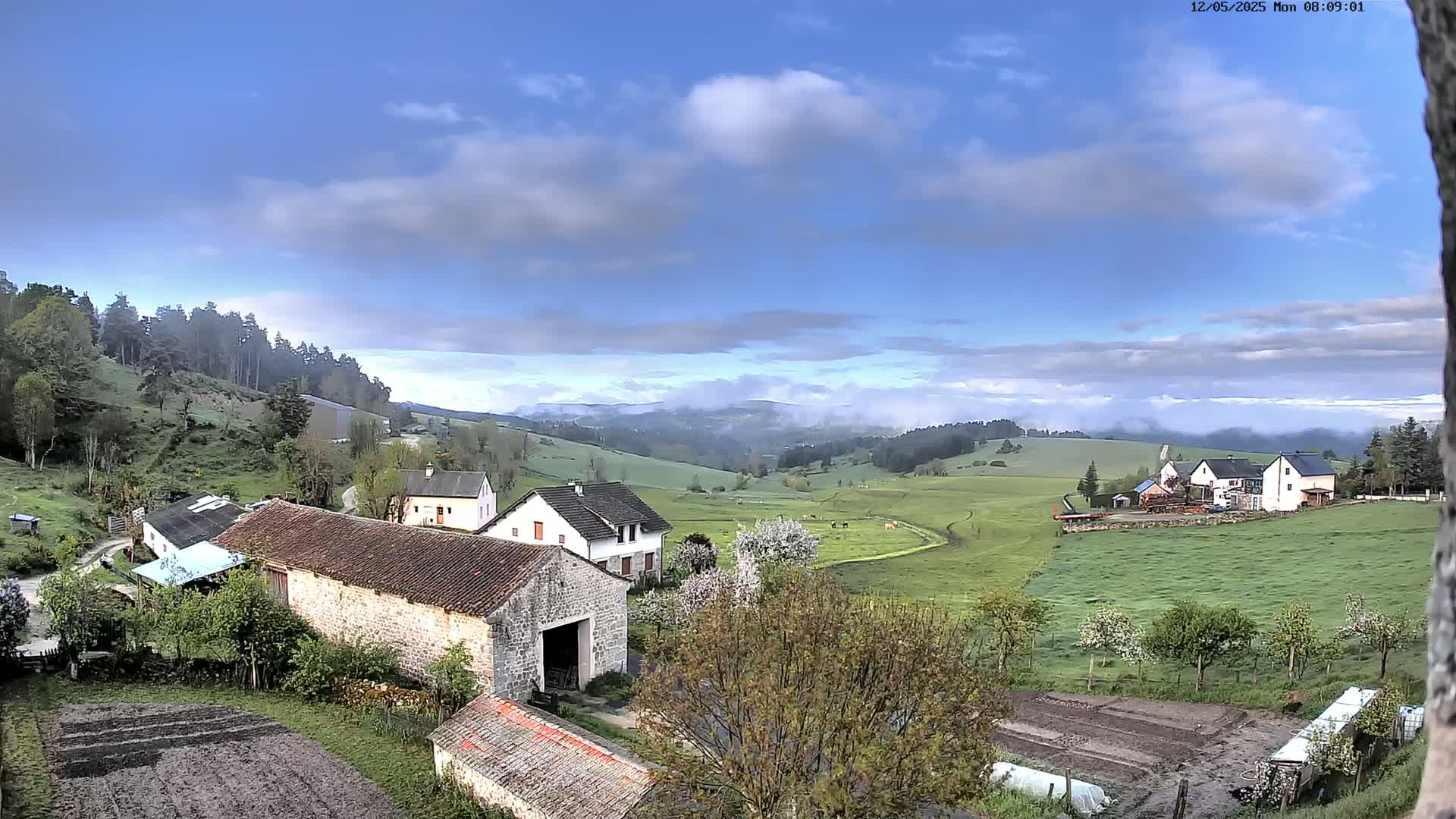 A partly cloudy sky overlooks a pastoral valley of green fields, stone farmhouses, and blossoming trees.