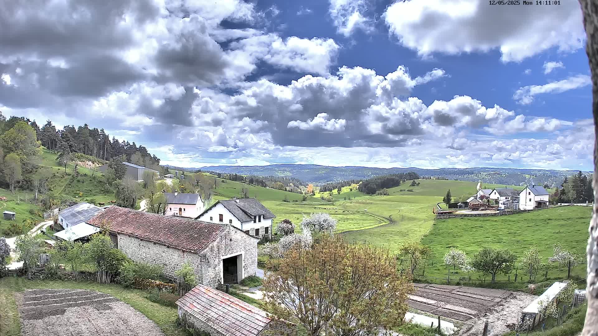 A partly cloudy day reveals a pastoral valley scene of stone farmhouses, barns, and tilled fields, nestled amid rolling green hills and distant forests.