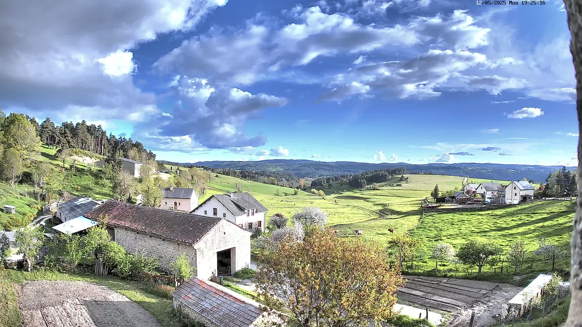 A partly sunny day reveals a rural landscape with stone farmhouses, green pastures, and rolling hills under a partly cloudy sky.