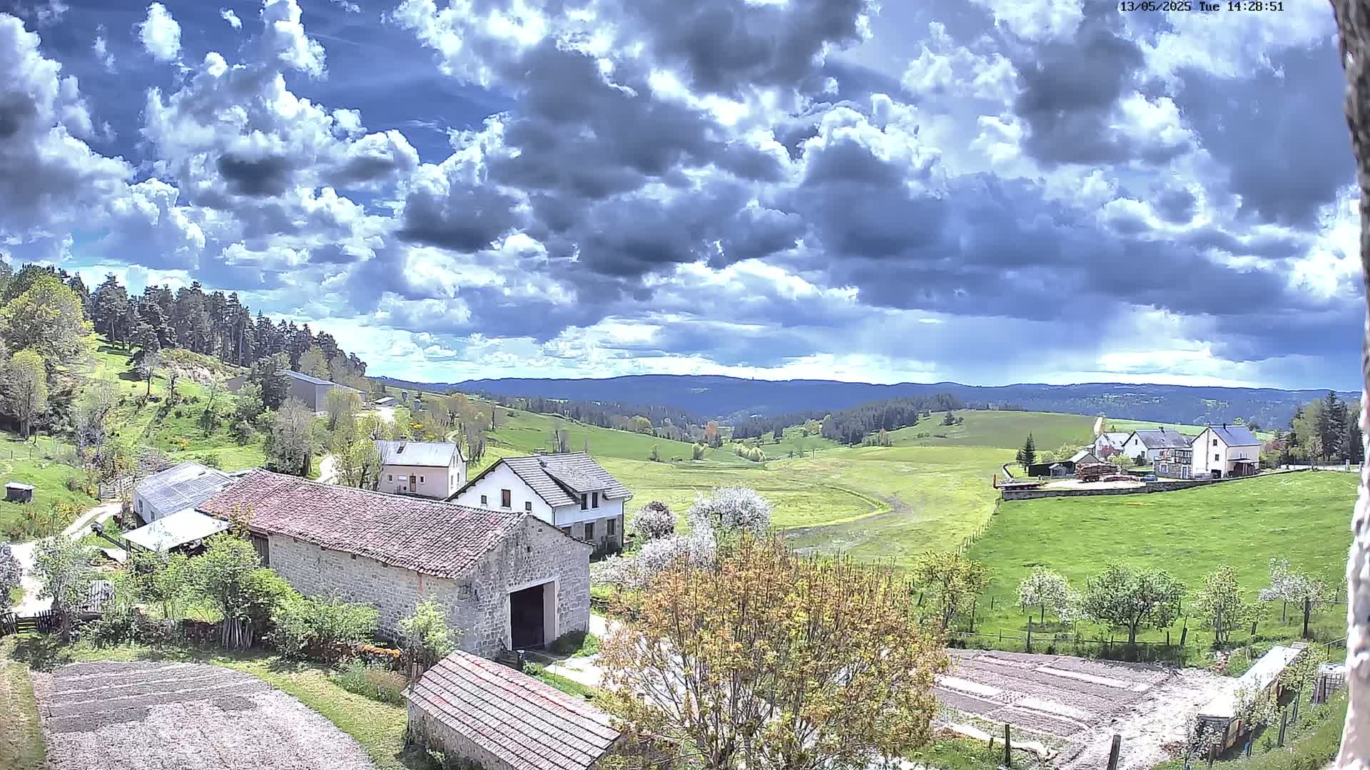 A partly cloudy day reveals a rural landscape of stone buildings, green fields, and a forest-covered mountain range in the distance.