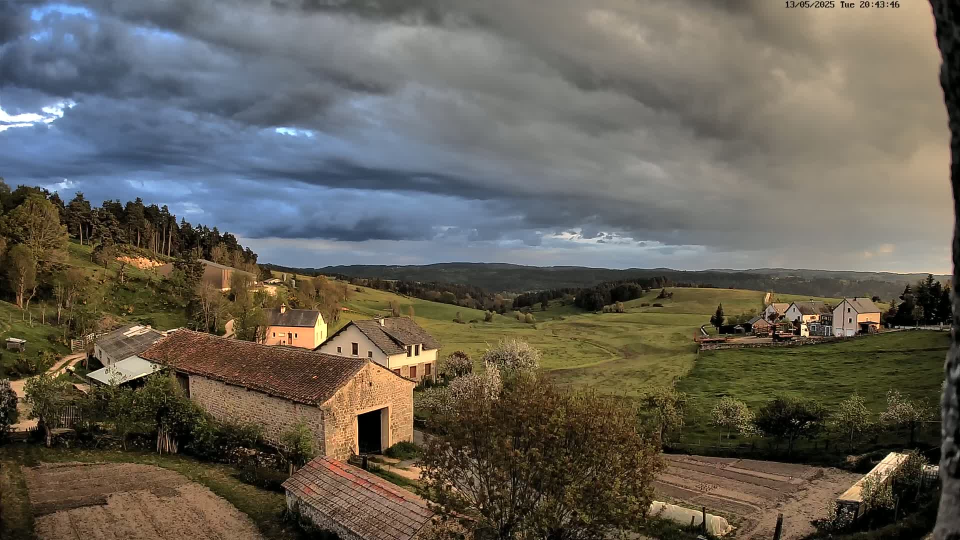 A rural landscape under a dark, cloudy sky shows several stone and wood buildings nestled among green hills and fields.