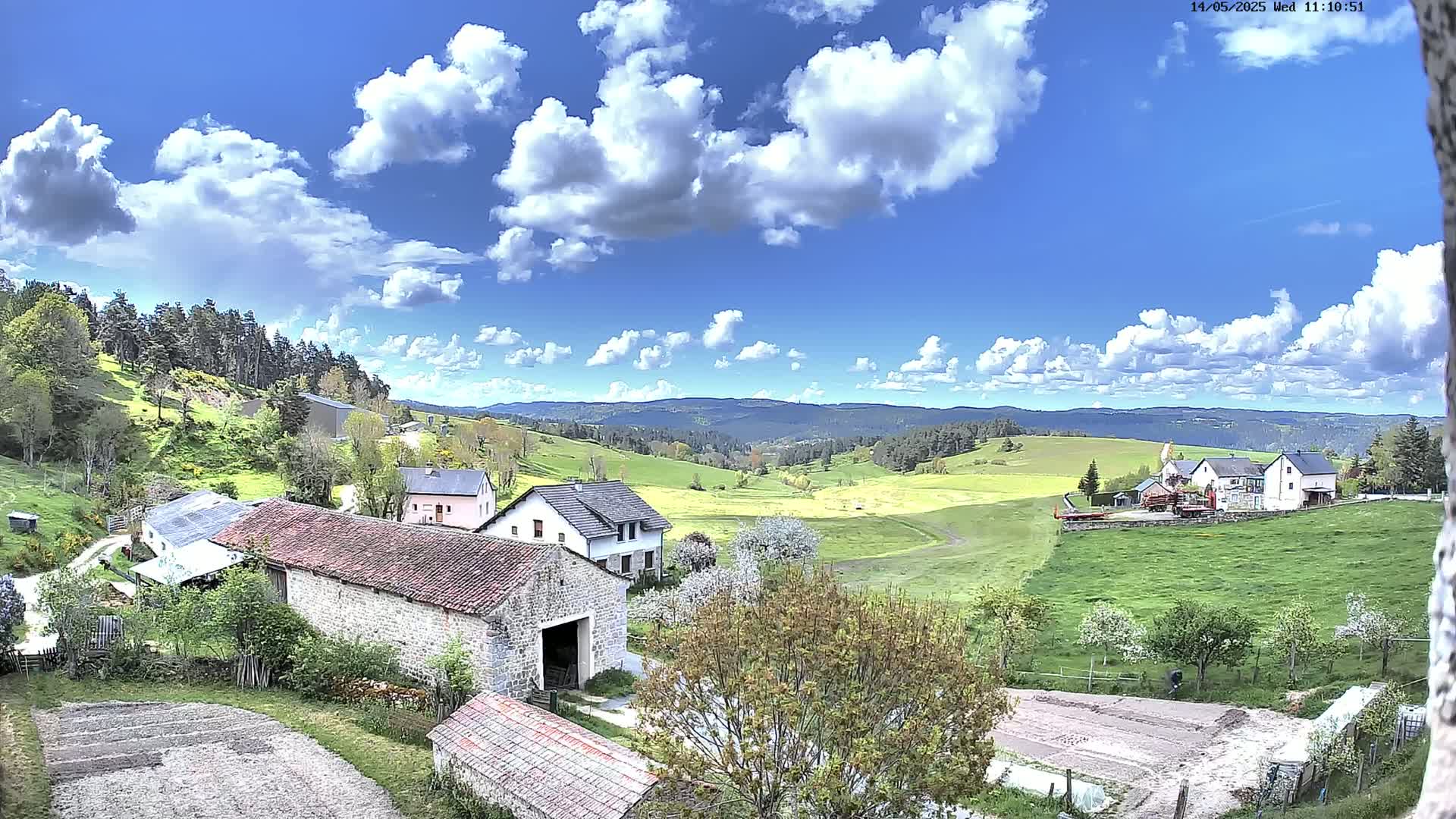 A partly cloudy blue sky overlooks a rural landscape of green fields, stone buildings, and blossoming trees.