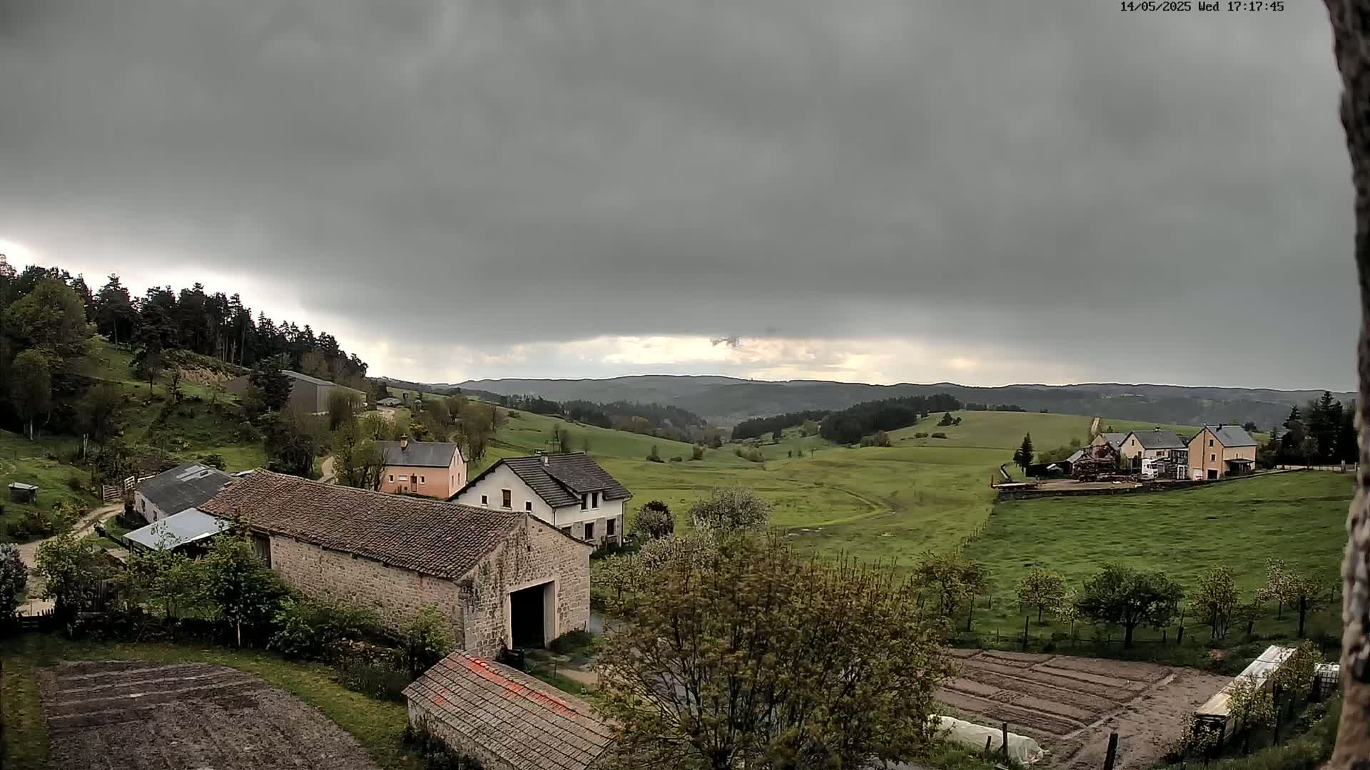 Under a gray, overcast sky, several stone farmhouses and barns sit nestled in a verdant valley, surrounded by rolling hills and a distant forest.