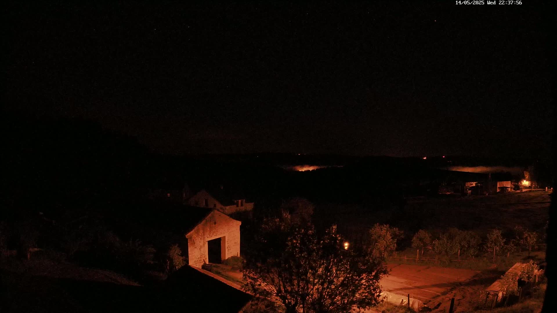 A nighttime view of stone buildings and trees, with a low-lying fog or mist in the distance under a clear, starry sky.