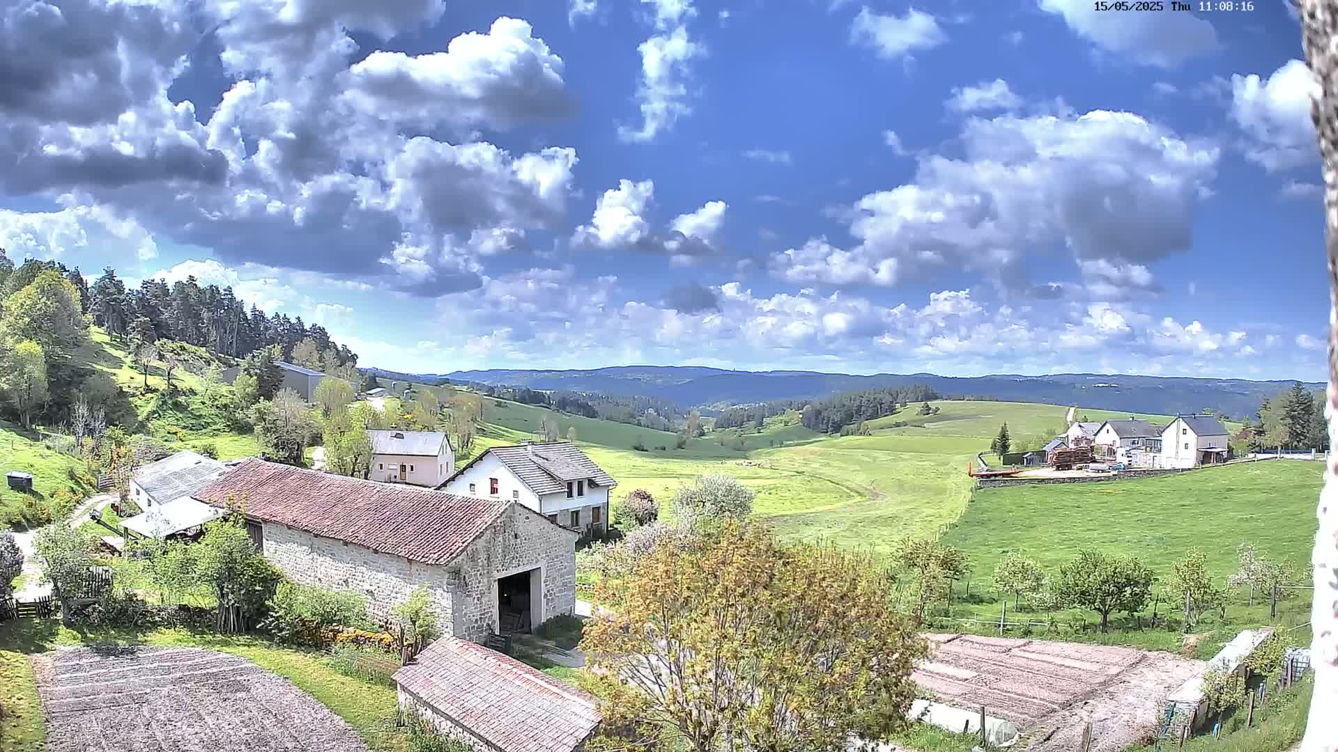 A partly cloudy sky overlooks a rural landscape of stone buildings, green pastures, and a distant forest-covered mountain range.