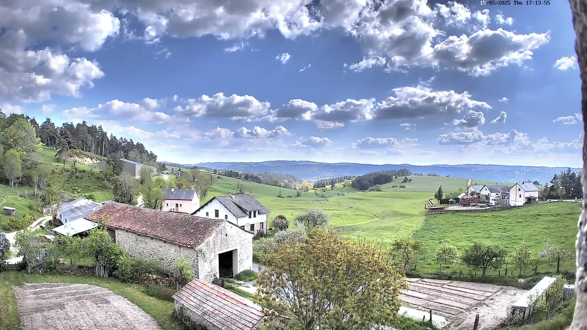 A partly cloudy sky overlooks a rural landscape of stone buildings, green pastures, and a distant forest under a mostly sunny sky.