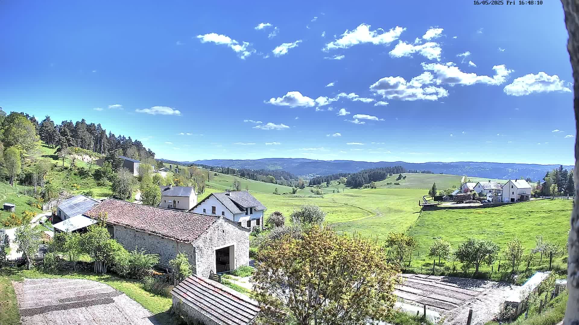 A sunny day with scattered clouds reveals a pastoral scene of stone farmhouses nestled in a green valley, overlooking rolling hills and distant forests.