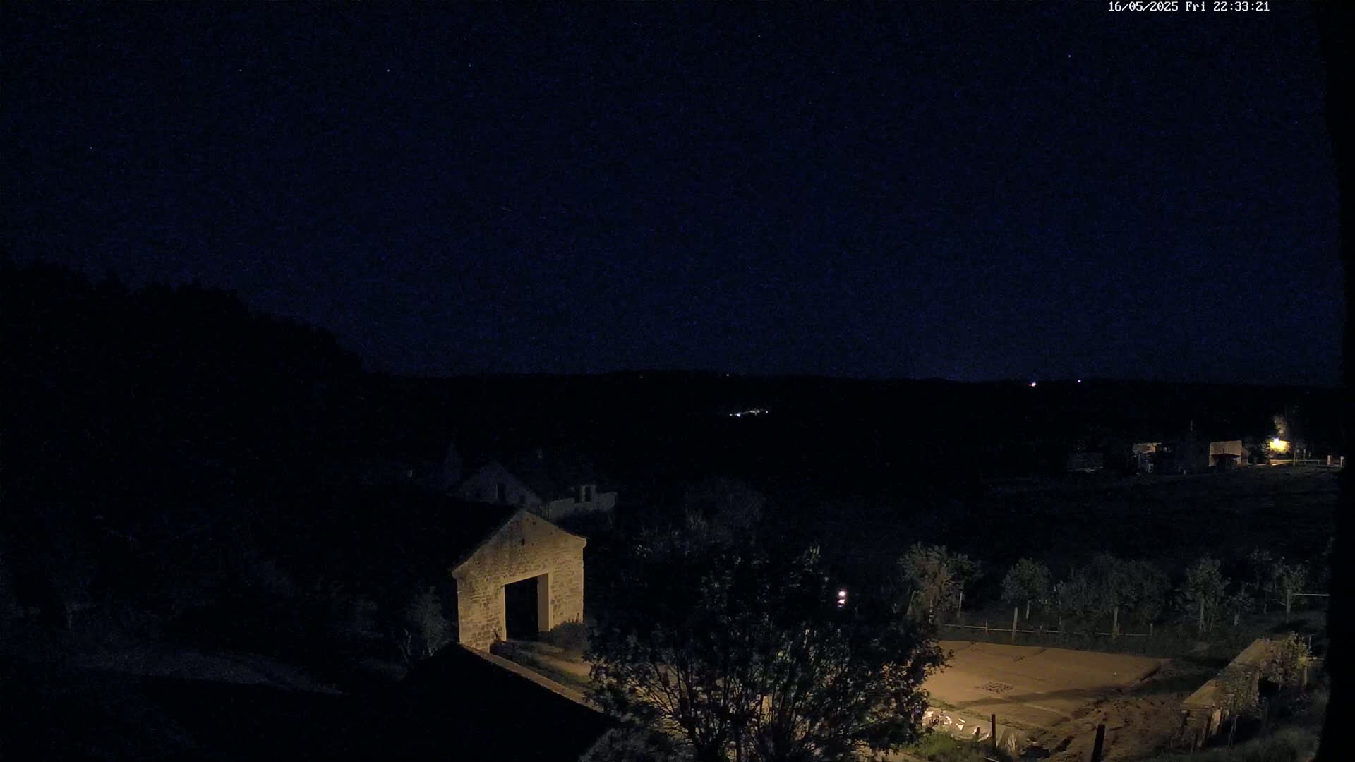 A stone structure is visible under a clear, dark night sky, with low-light illumination showing trees, a road, and other buildings in the distance.