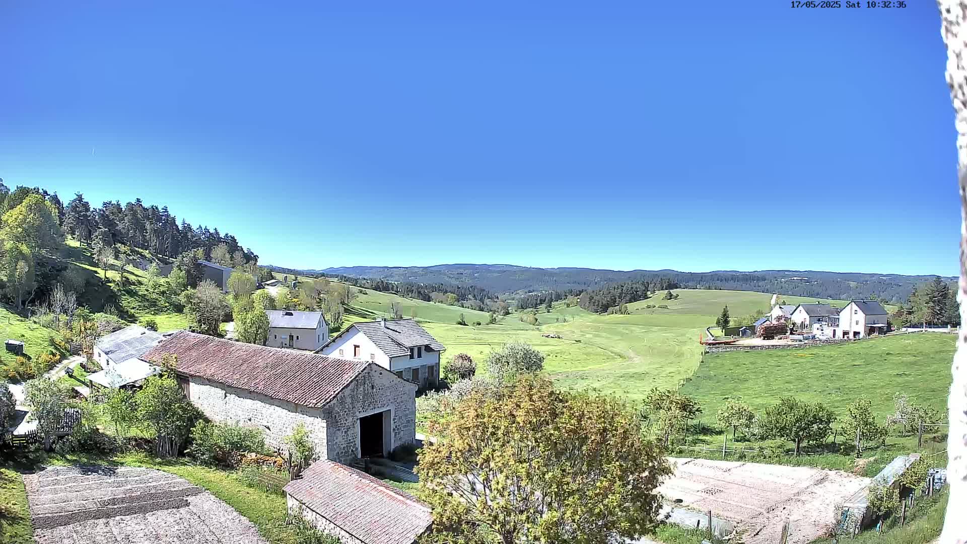 A sunny, rural landscape shows stone farm buildings nestled among green fields and rolling hills, with a forest visible in the distance.