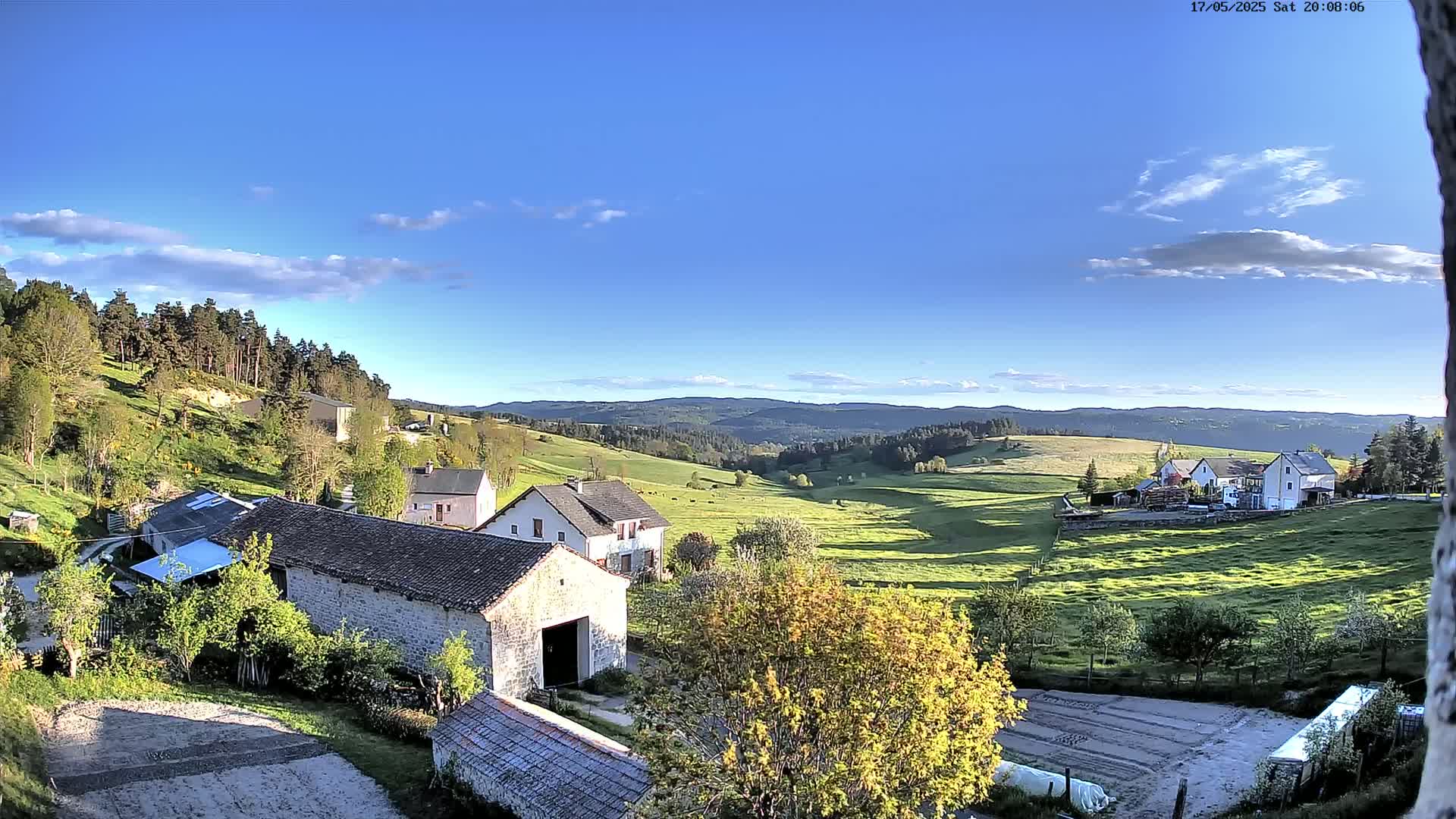 A rural scene of stone farmhouses and barns nestled in a green valley, under a mostly clear blue sky with a few scattered clouds.