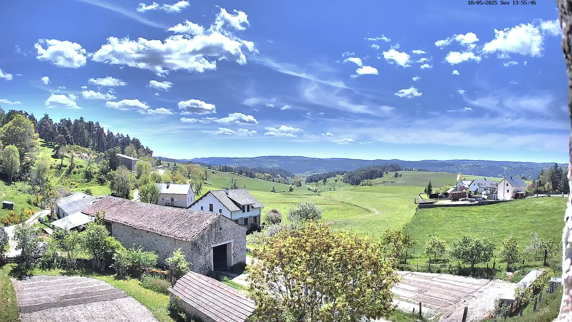 A sunny day with scattered clouds reveals a rural landscape of stone buildings, green pastures, and distant hills.