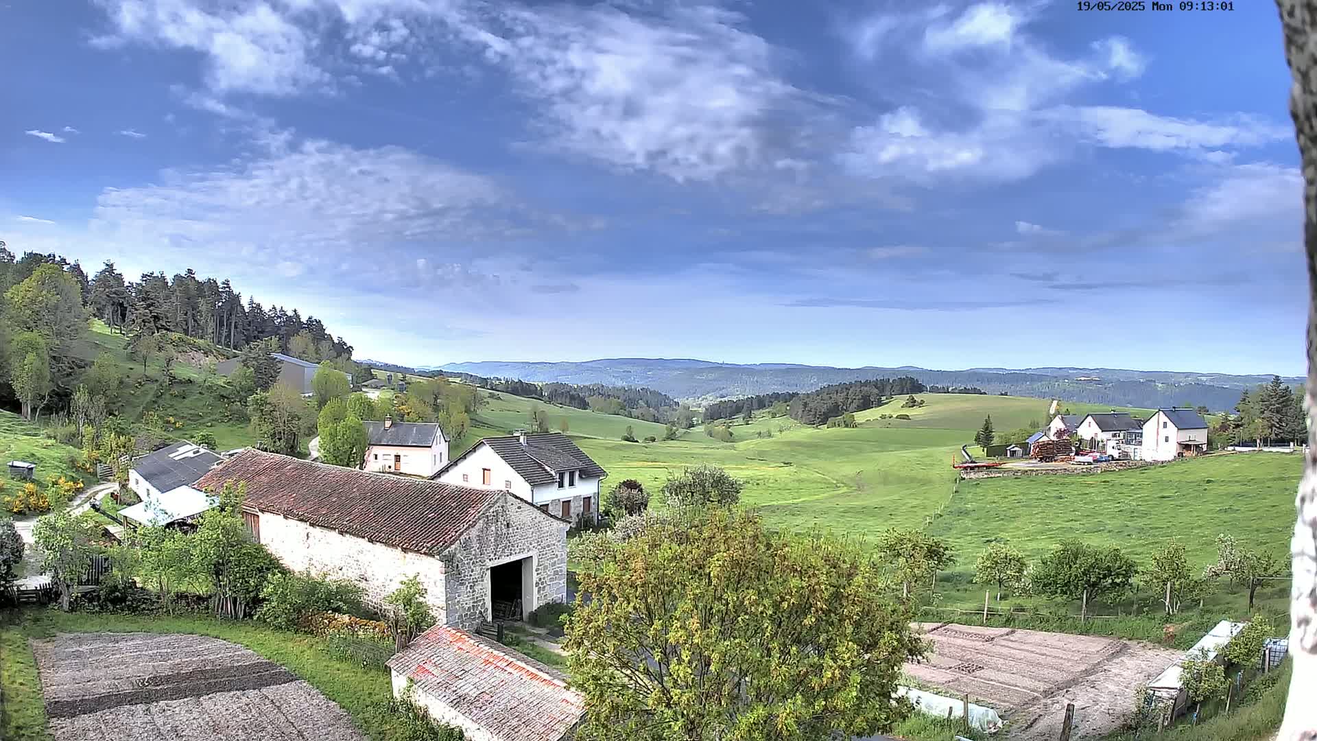 A partly cloudy sky overlooks a verdant valley dotted with farmhouses and rolling hills, with a forest in the distance.