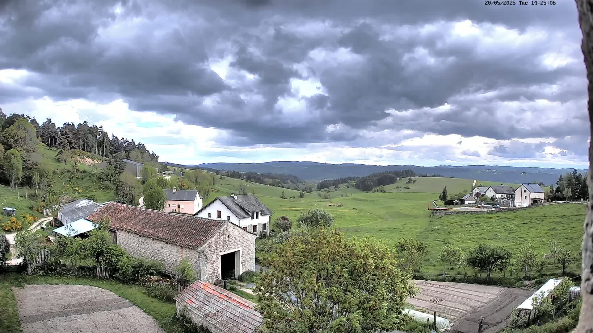 A rural landscape under a cloudy sky shows several stone buildings nestled among green fields and rolling hills, with a forest visible in the distance.