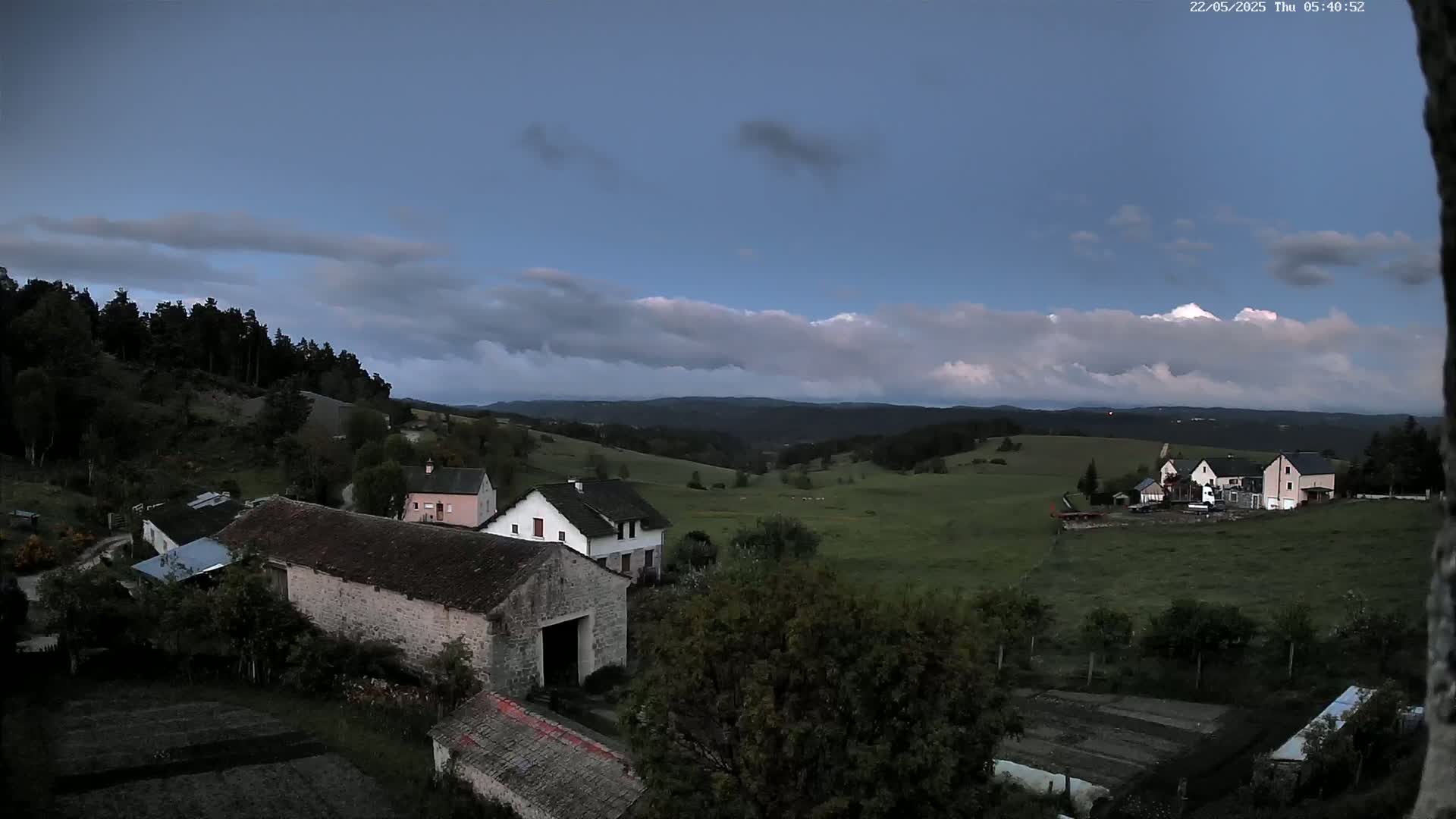 A rural landscape at twilight under a partly cloudy sky shows several houses and farm buildings nestled among rolling green hills and a dark forest.