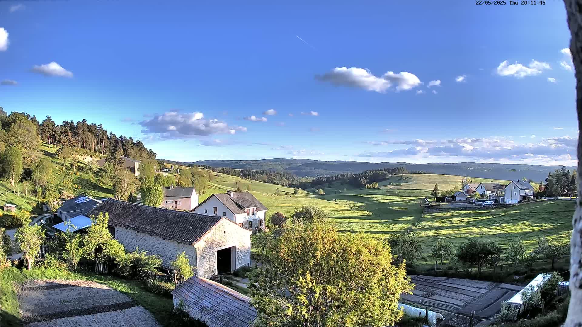 A sunny day with partly cloudy skies overlooks a rural valley with stone buildings, green fields, and a distant forest.