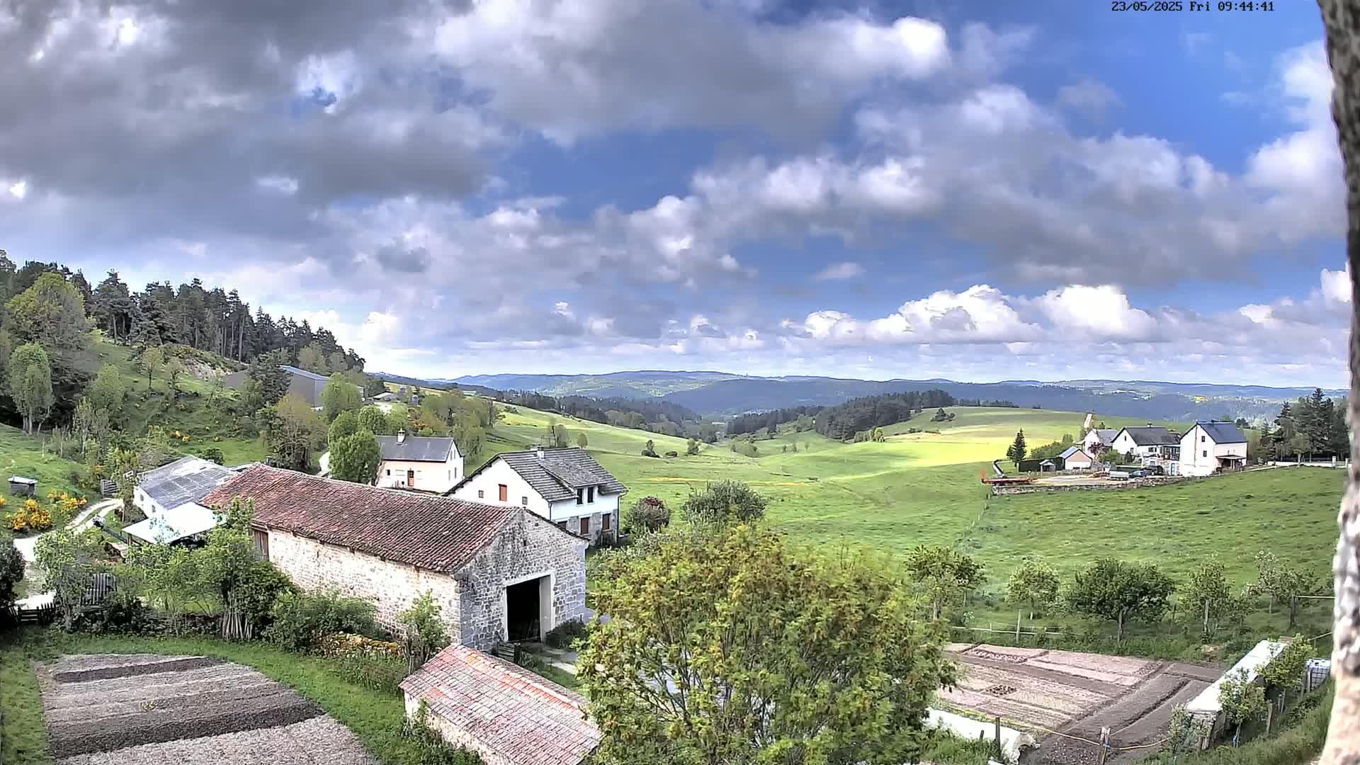 A partly cloudy sky overlooks a rural valley with several stone farmhouses, green pastures, and cultivated plots of land.