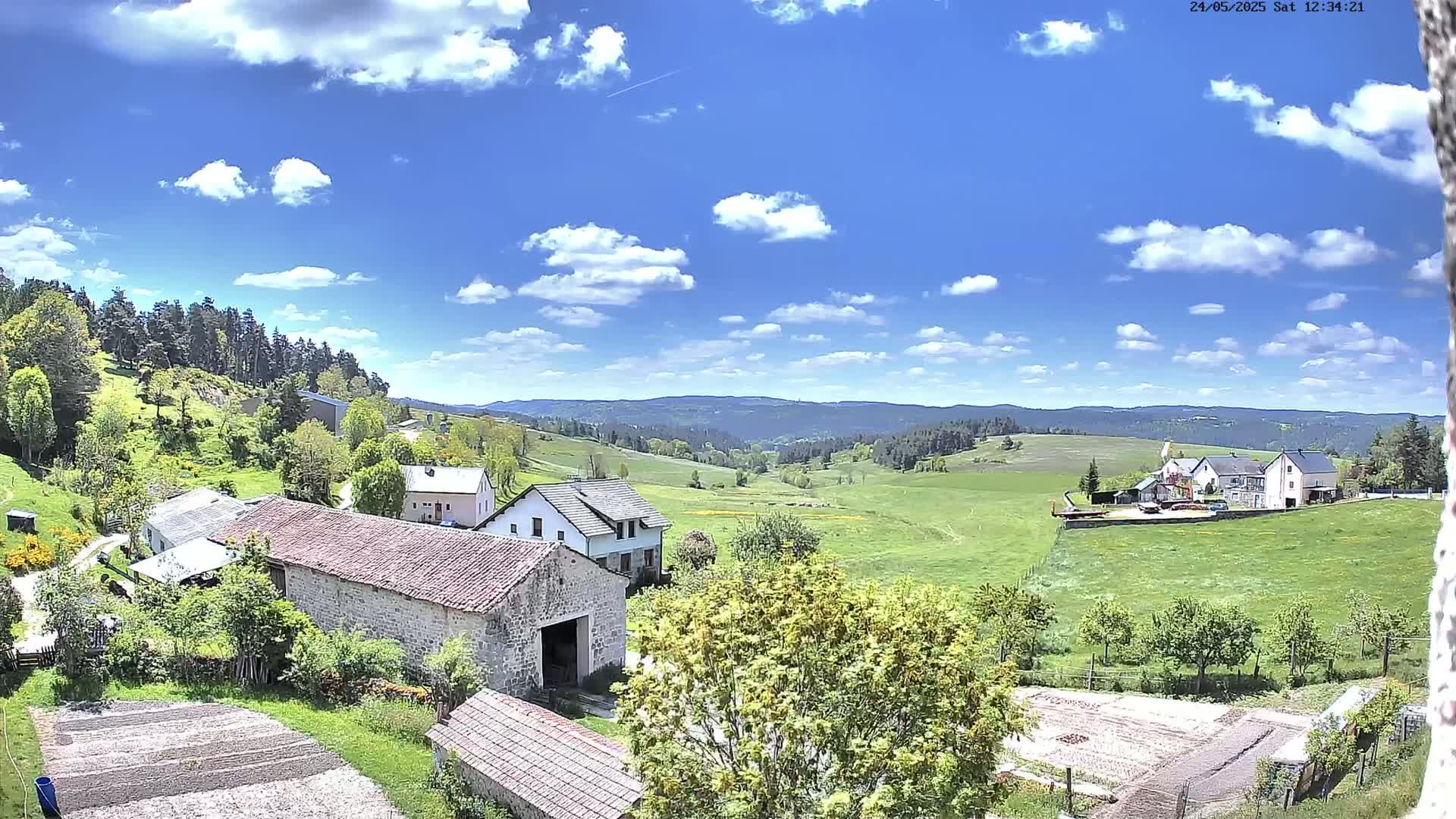 Stone farm buildings and houses are nestled in a verdant valley under a bright, partly cloudy sky.