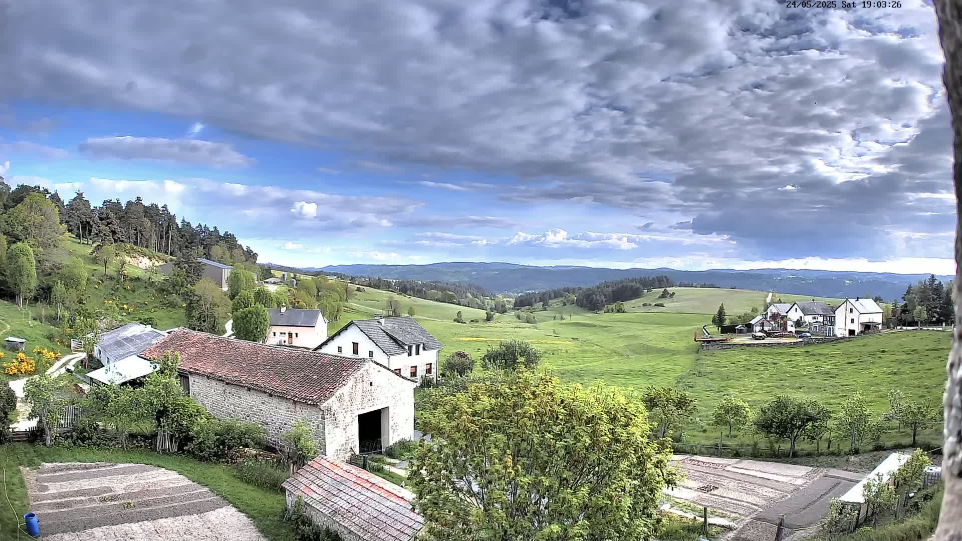 A partly cloudy sky overlooks a rural valley with several farmhouses and barns, green pastures, and forested hills in the distance.