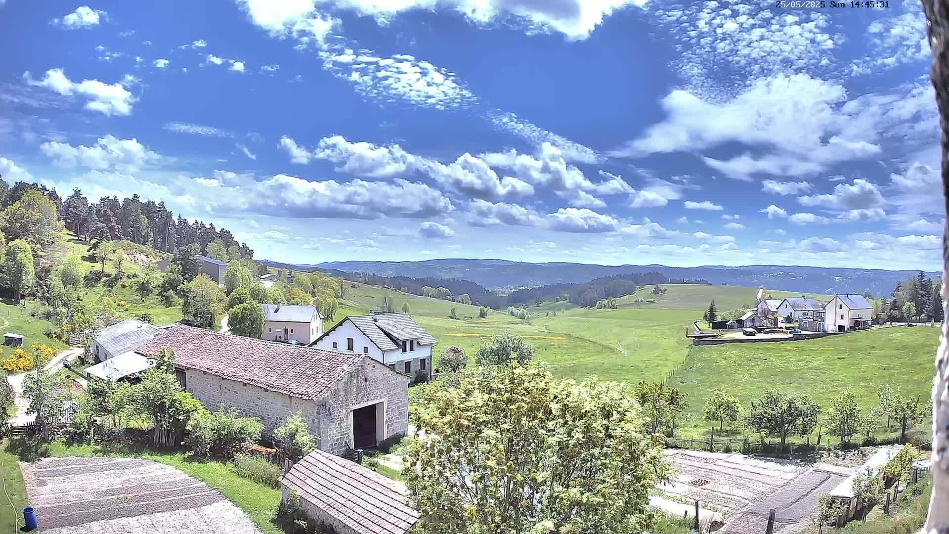 A sunny day with partly cloudy skies overlooks a rural landscape of stone farm buildings, green pastures, and distant rolling hills.