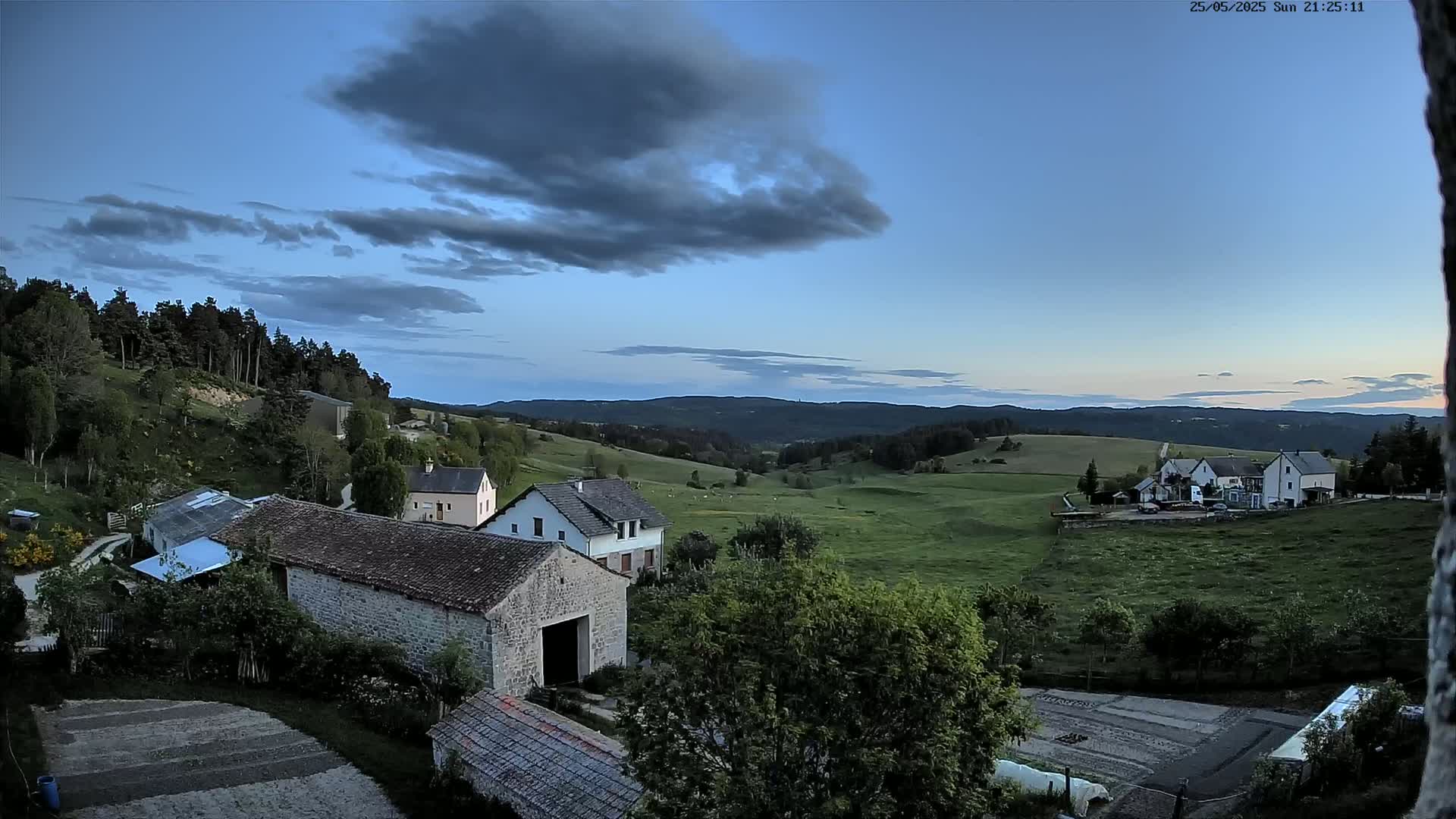 A sprawling rural landscape at dusk, under a partly cloudy sky, features several stone buildings nestled amongst green fields and a forest-covered hill.