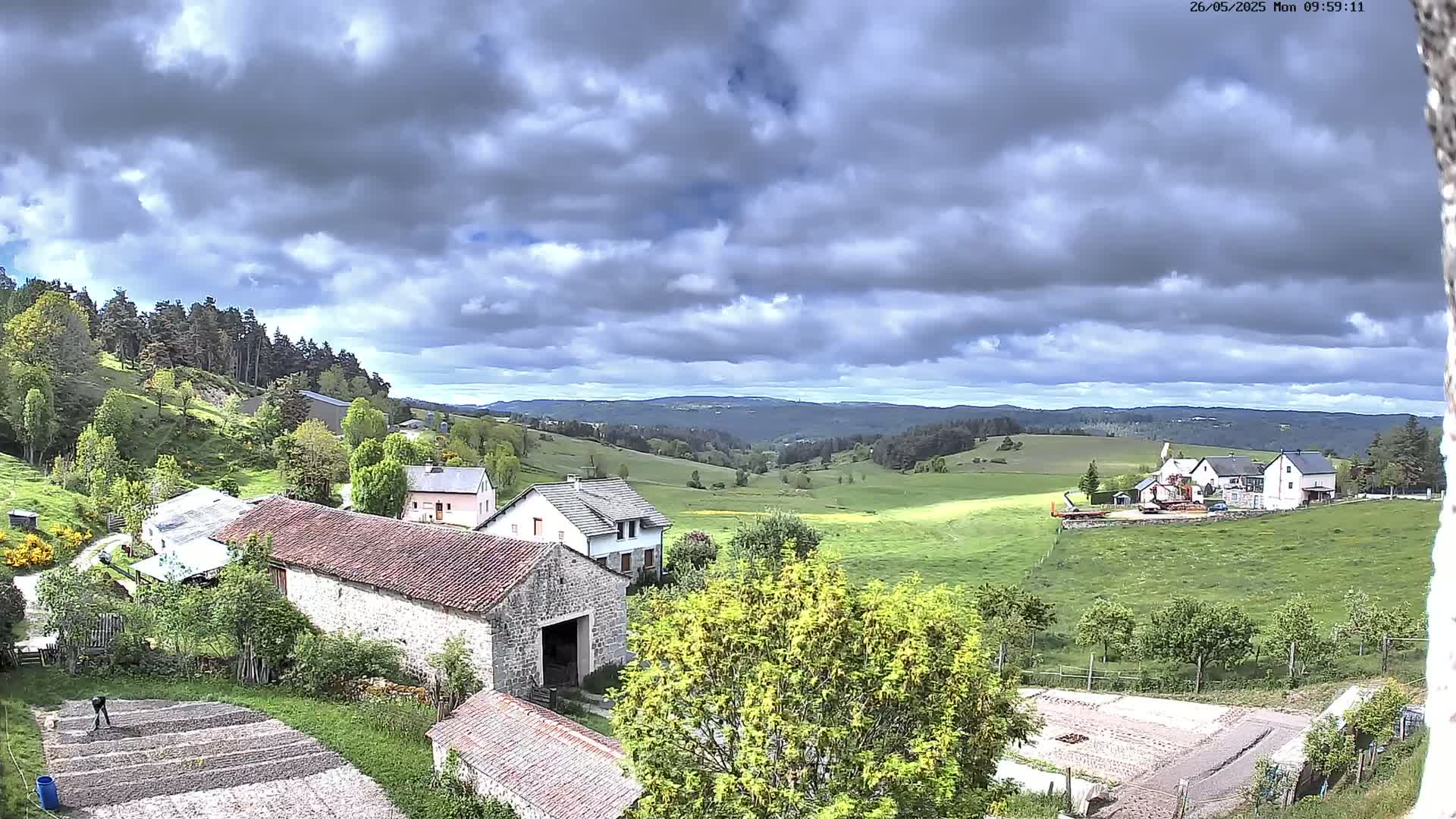 A rural landscape under a partly cloudy sky shows several stone buildings and houses nestled amongst green fields and hills, with a forest in the background.