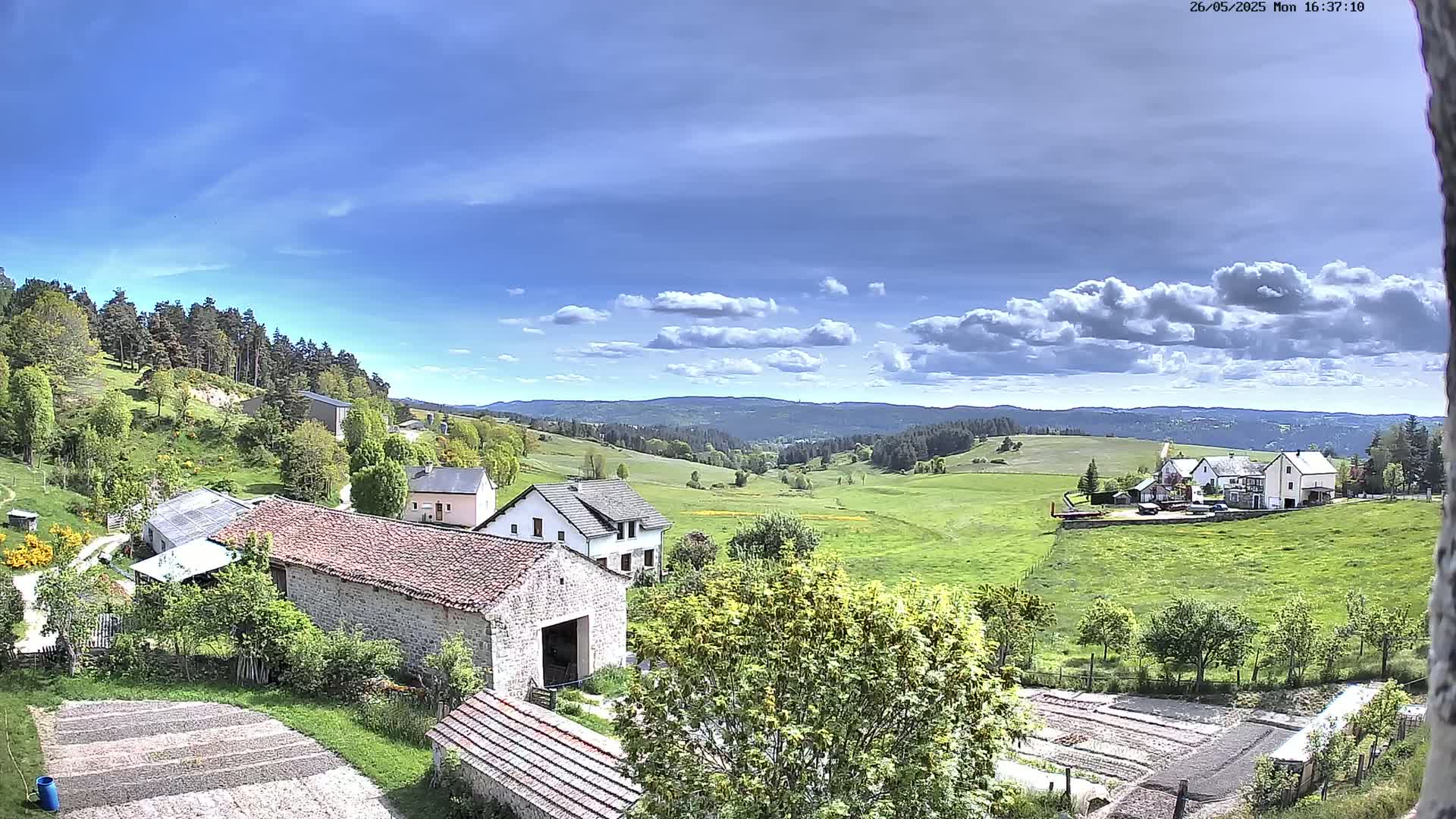 A partly sunny day reveals a picturesque rural landscape of rolling green hills, stone farmhouses, and a forest-covered mountain in the distance.