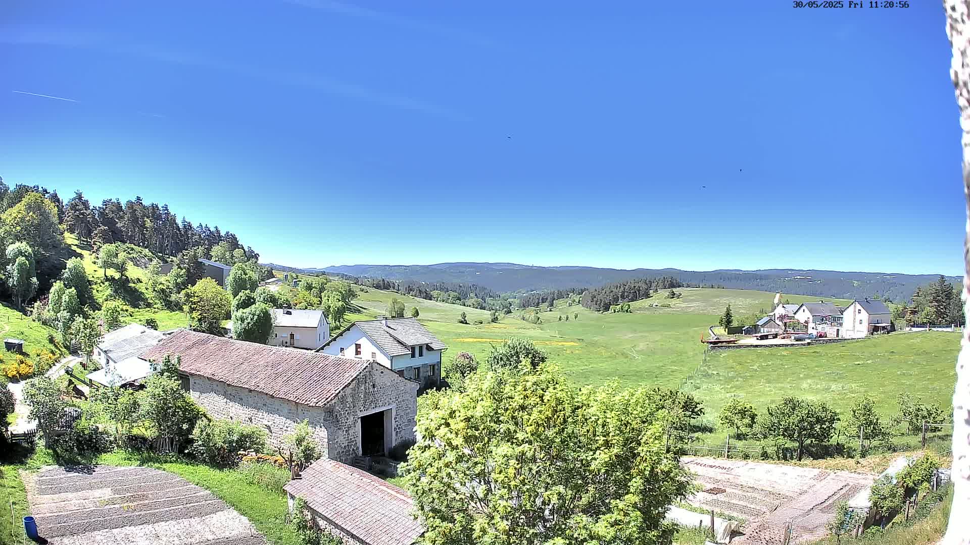 A sunny, clear day reveals a rural landscape with stone buildings, green fields, and a distant forested mountain range.