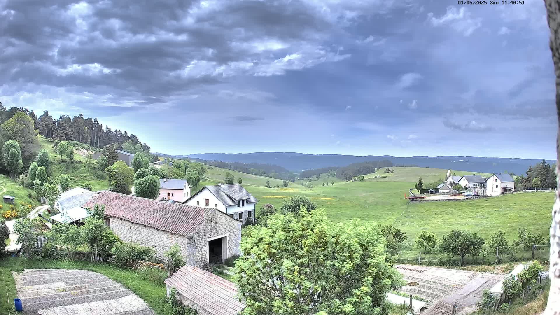 A rural scene under a partly cloudy sky shows several stone buildings, green fields, and a forest in the background.