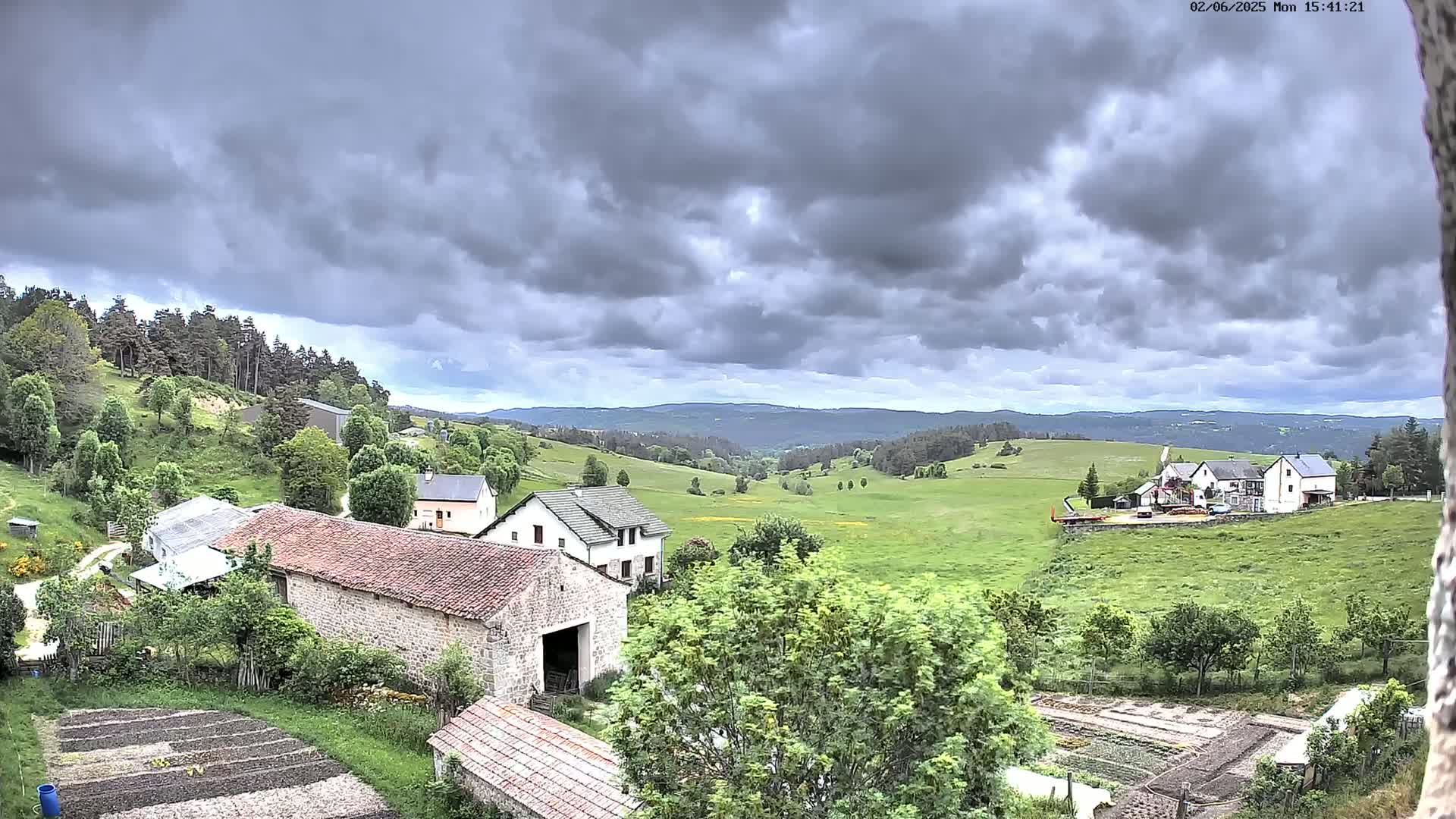 A rural landscape under a heavily clouded sky shows several farm buildings nestled among green fields and gardens, with a distant view of rolling hills and forests.