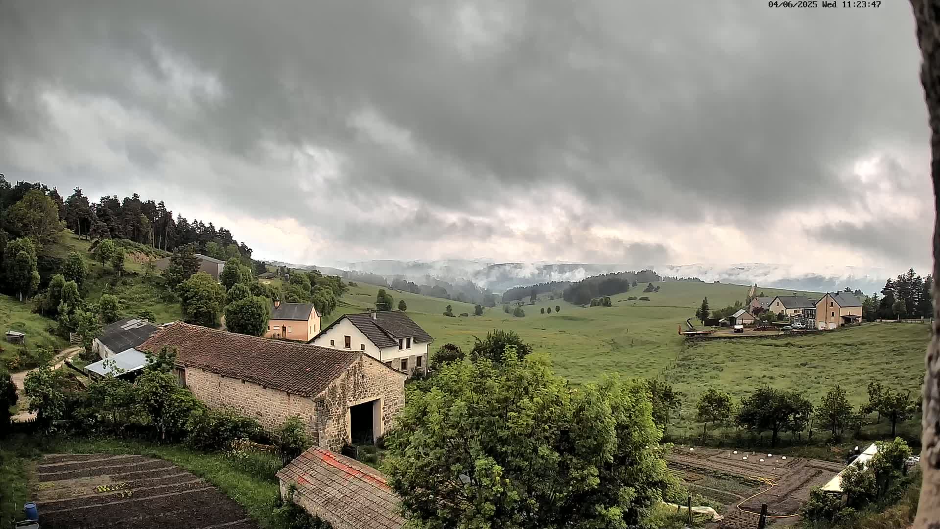 A rural landscape under a cloudy sky shows several stone buildings, green fields, and a line of trees in the distance.
