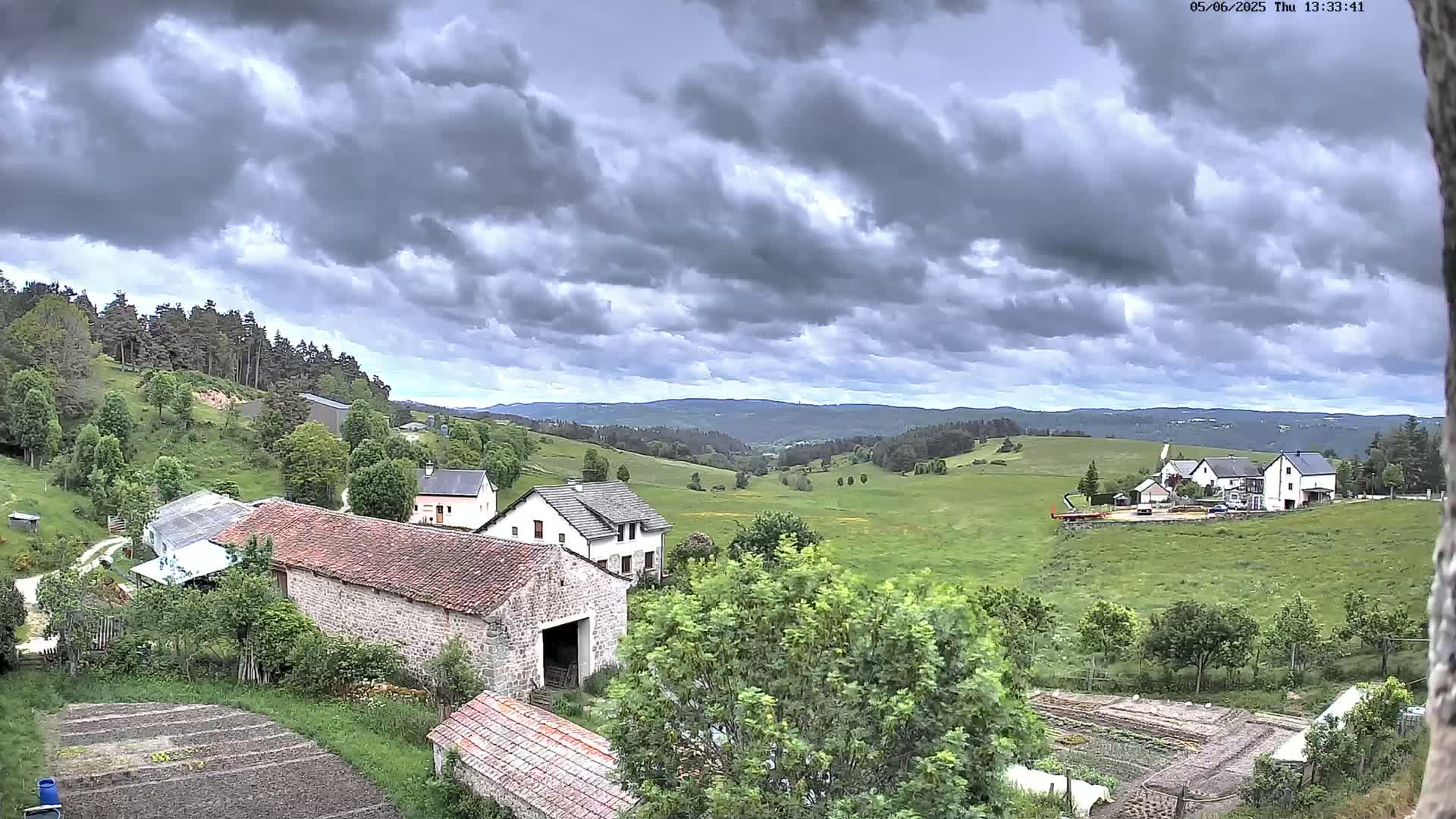 A rural landscape under a cloudy sky shows several buildings nestled amongst green hills and fields, some with cultivated gardens nearby.